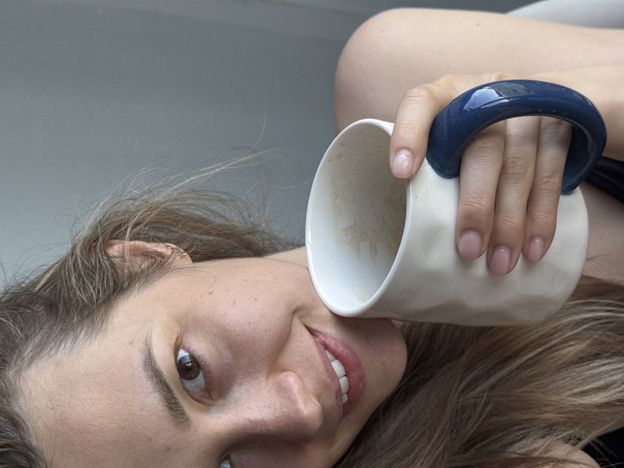 A woman with light brown hair and fair skin is smiling while holding a white ceramic mug with a dark blue handle. The mug appears partially empty, containing some residue inside. The woman's face is in the lower left portion of the frame, and the mug is positioned to her right. The background is a plain, light gray color. The image does not contain any information that would indicate it was taken in Chaikovsky, Russia, or any other specific location.
