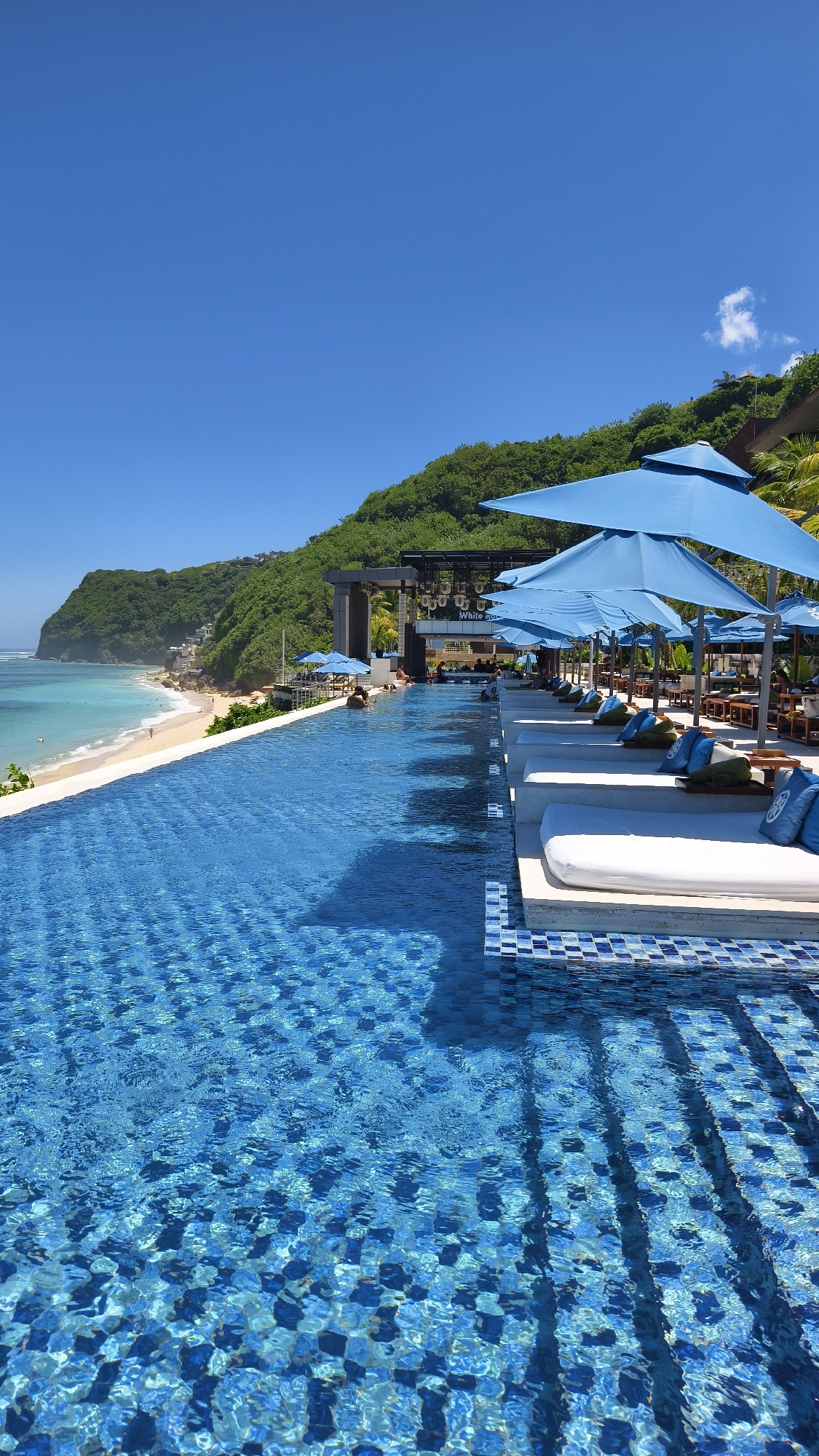 The image shows a resort swimming pool overlooking a beach in Kuta Selatan, Indonesia. The pool is tiled with blue and white mosaics. Along the pool's edge are lounge chairs under blue umbrellas. A beachfront building is visible behind the pool. The beach is sandy with turquoise water, and a green hillside rises in the background. The sky is clear and blue.