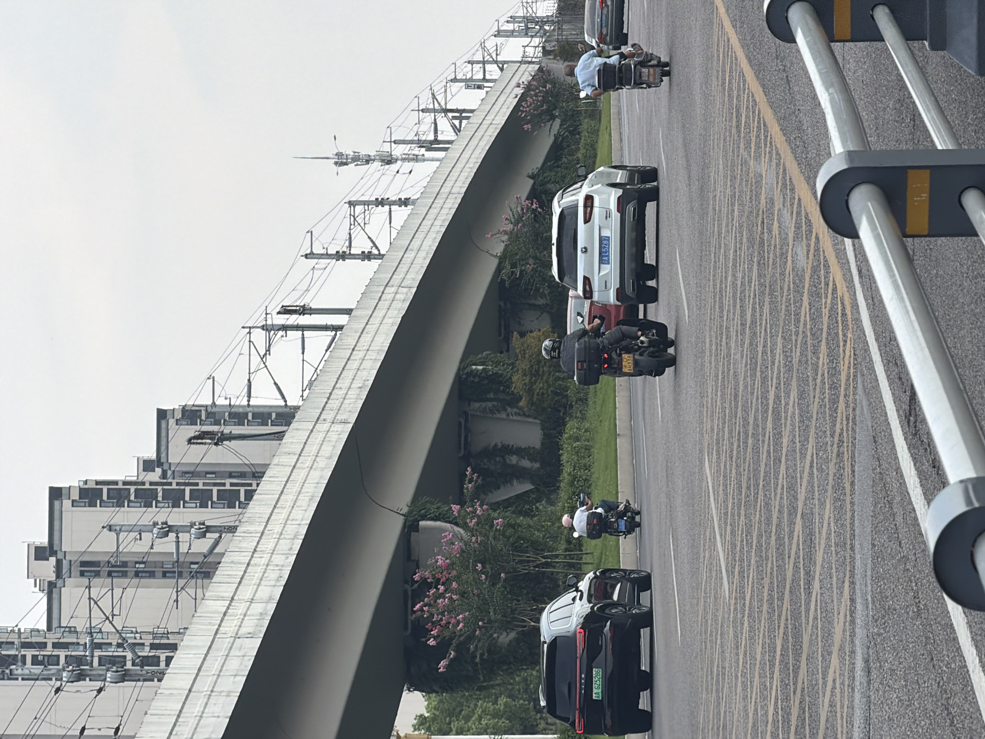 In Jiangjin, China, this image captures an urban street scene with elevated infrastructure and vehicles. A large, concrete overpass stretches across the frame, supporting overhead power lines and complex electrical infrastructure, characteristic of urban transportation networks. Beneath the overpass, several vehicles are visible on a multi-lane road. A black SUV, identified by its license plate AA GZ5286, travels in the left lane. To its right, a white SUV with license plate AL5287 is also in motion. Several individuals are riding motorcycles. One motorcycle rider, wearing a helmet and black clothing, is in the foreground. Another motorcycle rider, wearing a light blue shirt, is further to the right. In the distance, additional vehicles are present, including a silver sedan. The surrounding environment features trees and greenery, with pink flowering bushes visible beneath the overpass. The sky is overcast, suggesting a cloudy day. The road surface is asphalt, marked with yellow and white lines, including a large cross-hatched yellow area, possibly indicating a no-parking zone or a pedestrian crossing. Metal guardrails are present along the side of the road. Residential buildings with multiple stories are visible in the background, behind the elevated structure.