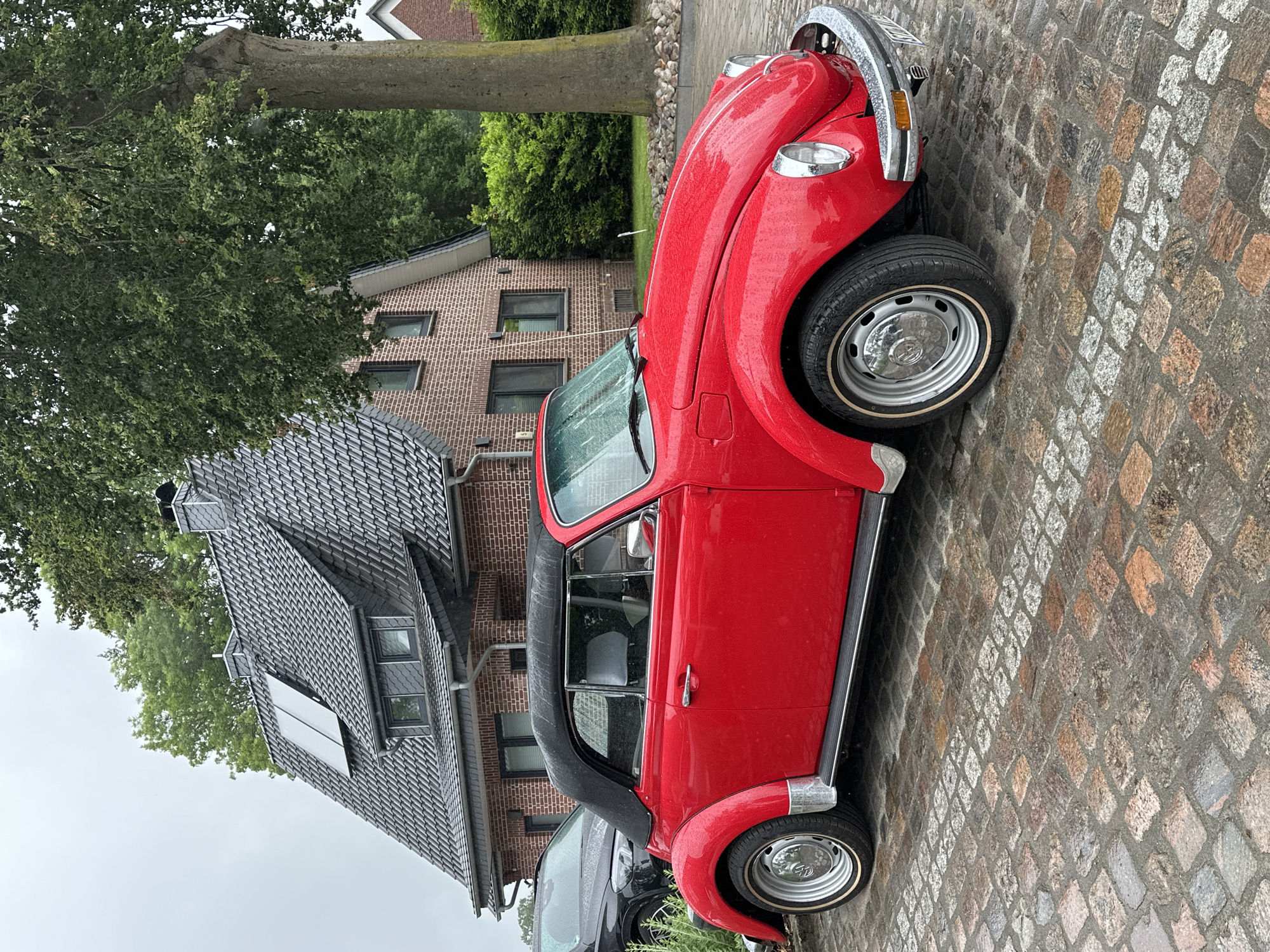 A bright red classic Volkswagen Beetle convertible is parked on a wet cobblestone driveway in Deinste, Germany. The vehicle is covered in visible raindrops, and its black soft-top is raised. Chrome bumpers, hubcaps, side trim, and door handles are prominent, and the windshield wipers are in an upright position. Behind the red Beetle and to its left, the front section of a dark-colored modern SUV is partially visible, also appearing wet. The driveway is paved with multi-toned granite cobblestones, which are slick with moisture, and a distinct diagonal pattern of lighter-colored stones is visible in the foreground. In the background, a two-story house with a red-brown brick facade and a steeply pitched dark grey tiled roof is visible. The roof features two solar panels, multiple dormer windows with dark frames, and two dark grey chimneys. A large, mature tree with a thick trunk and dense green foliage stands to the right of the house, partially obscuring a distant structure. Additional green trees and shrubs are present throughout the background. The sky is overcast, indicative of rainy weather. No individuals are present in the image.
