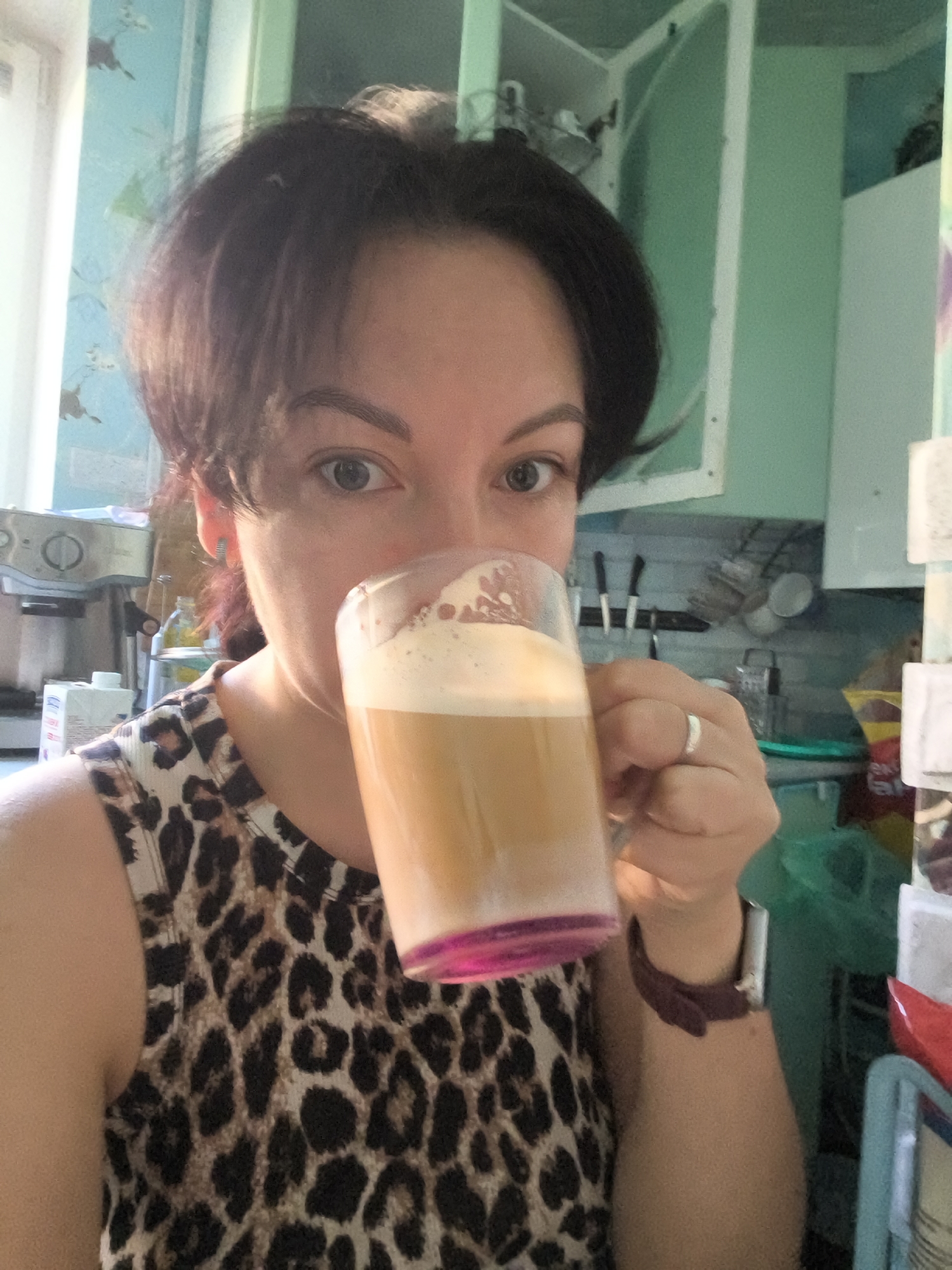 The image shows a selfie of a woman with short, dark hair, holding a glass of coffee with foam and a pink-colored bottom, to her lips. She is wearing a leopard-print sleeveless top and a ring on her left hand. The background appears to be a kitchen, with cabinets and what looks like a coffee machine. There is no information to suggest the location is in Orenburg, Russia.