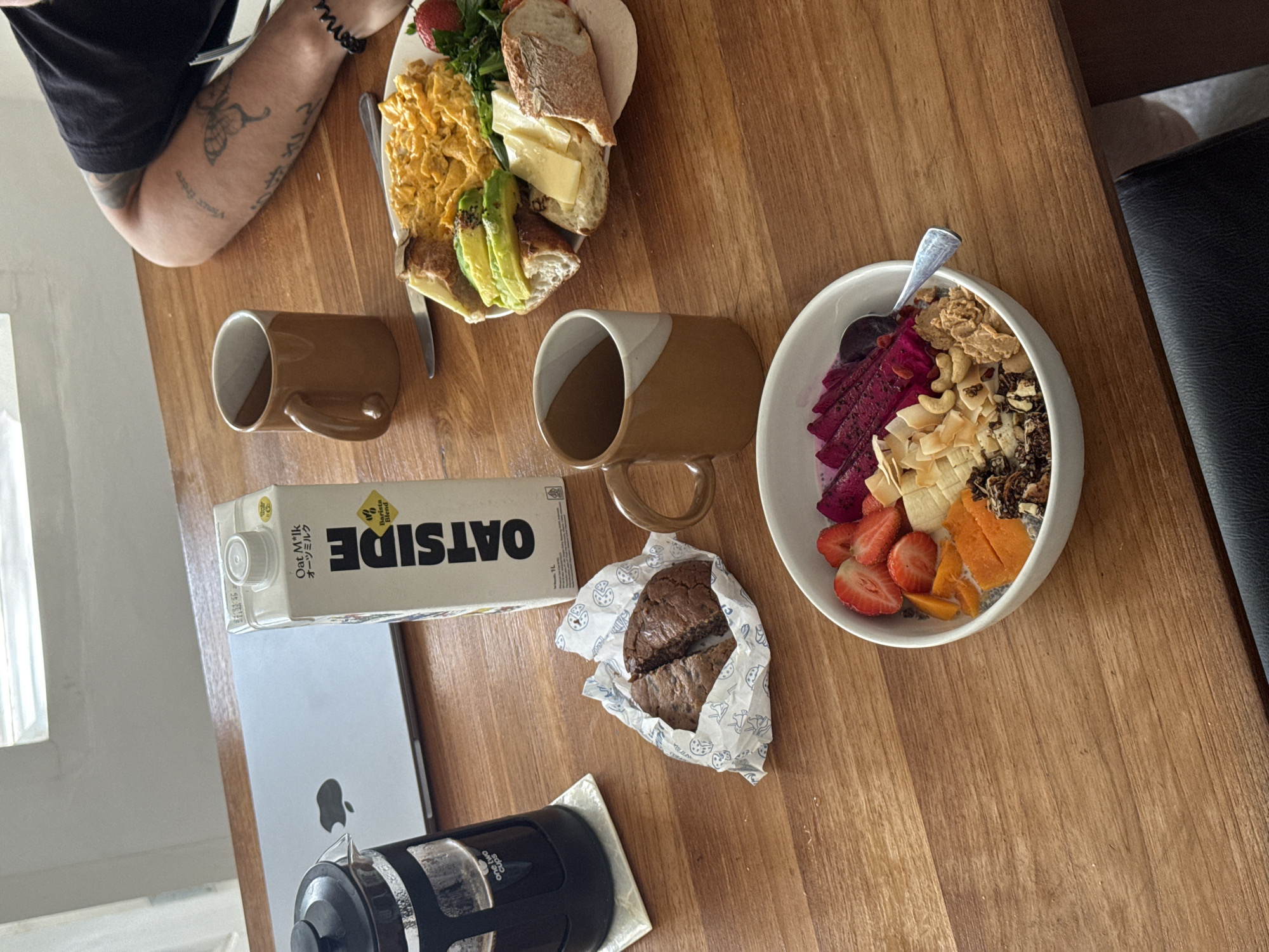 A breakfast spread is laid out on a wooden table in an indoor setting. In the foreground, a white bowl overflows with a colorful acai bowl topped with sliced strawberries, dragon fruit, mango, bananas, granola, cashews, and coconut flakes. To the right of the acai bowl is a white plate containing a breakfast of scrambled eggs, avocado slices on toast, a slice of bread with cheese, and a strawberry. Two brown ceramic mugs are placed on the table. To the left, a black French press sits on a coaster next to an open laptop with an Apple logo. A carton of Oat M*lk is positioned between the laptop and the mugs. A crumpled wrapper holding two halves of a chocolate cookie is placed in front of the French press. A person's tattooed arm and shoulder are visible on the right side of the image. The scene appears to be in North Kuta, Indonesia.