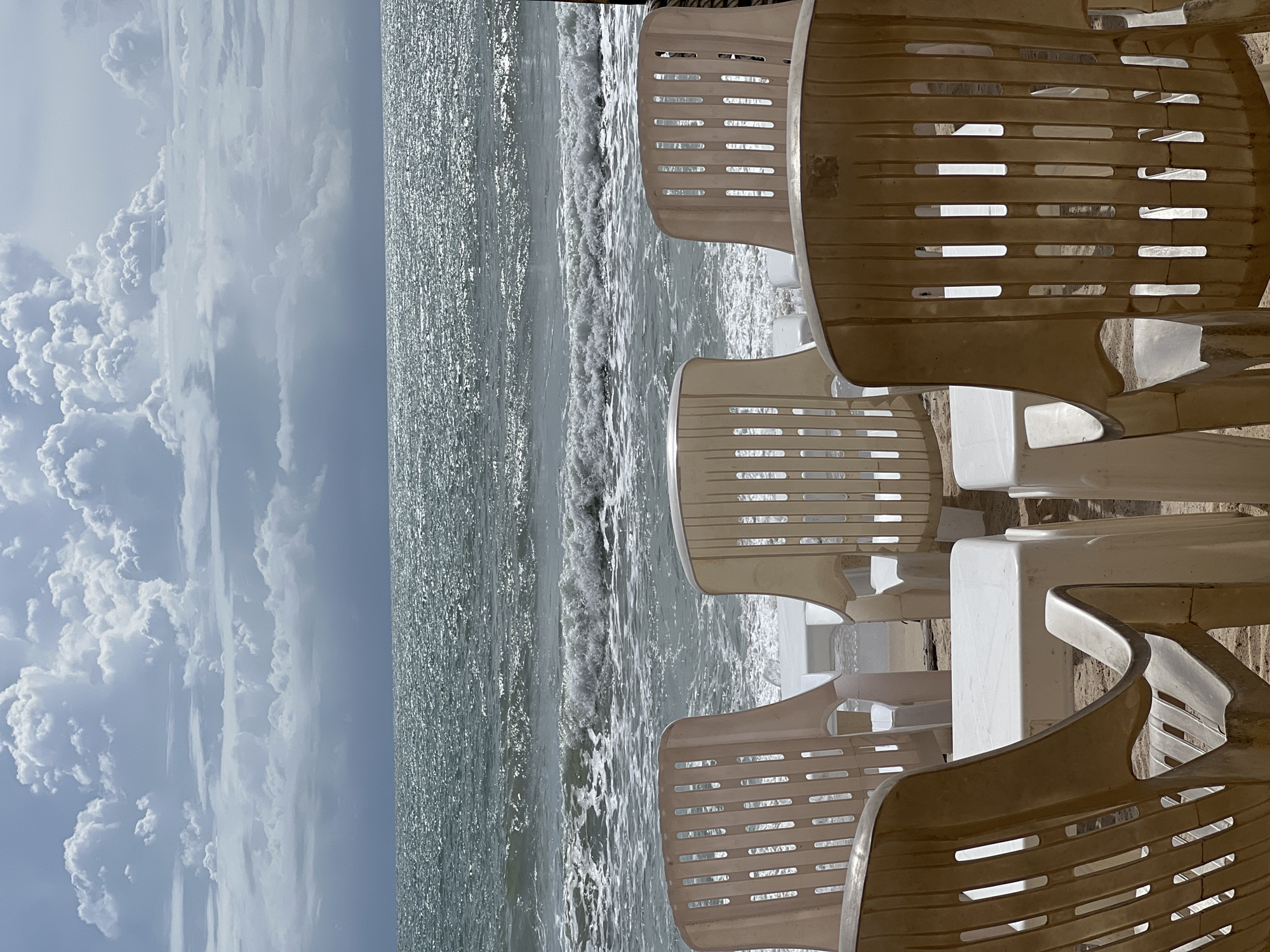 This detailed image captures an outdoor scene at a beach in Unknown, Unknown, under a dynamic cloudy sky. In the immediate foreground, five plastic chairs and two small plastic tables are arranged on sandy ground, facing the ocean. Three chairs, viewed from behind, are prominently positioned with two tables between them. These chairs are light beige or tan, with vertical slat designs on their backs and armrests; one chair to the far right appears slightly darker or more discolored. Further back, two lighter-colored plastic chairs are visible, flanking the central arrangement, also facing the sea. The chairs and tables are unoccupied.
The midground is dominated by the ocean, where moderate waves with white foam break near the sandy edge. The water's surface, particularly further from the shore, exhibits strong glints of sunlight, creating a shimmering effect across the expanse of the sea. The horizon line clearly separates the sparkling ocean from a heavily clouded sky.
The background features a vast expanse of sky filled with large, towering cumulus clouds. These clouds display varying shades of white and grey, suggesting depth and thickness, with brighter areas indicating direct sunlight and darker areas possibly indicating denser cloud cover or shadows. No individuals or man-made structures beyond the furniture are visible.