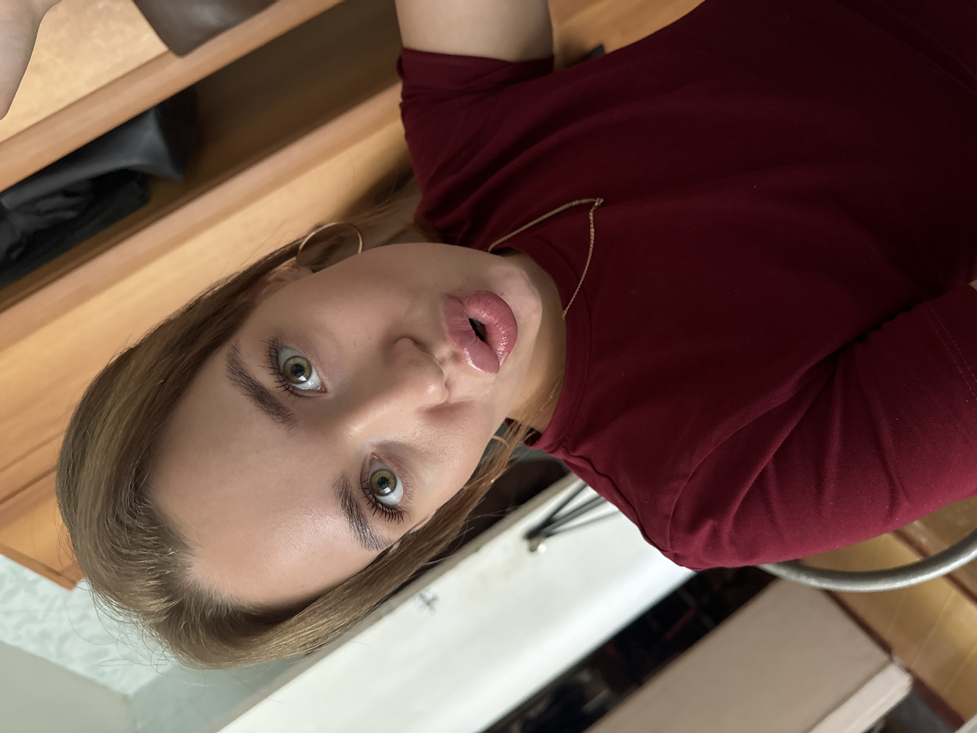 A young woman with light brown hair and green eyes is captured in a close-up, slightly tilted shot. She is wearing a dark red, long-sleeved shirt and a delicate gold necklace. Her lips are parted, and she appears to be looking directly at the camera with a neutral expression. Large hoop earrings are visible on her ears. The background suggests an indoor setting with wooden furniture and what looks like a white appliance or countertop. The lighting is soft, and no specific time of day or weather conditions can be determined. The image does not contain any visible text. The location is contextually given as Chaikovsky, Russia.