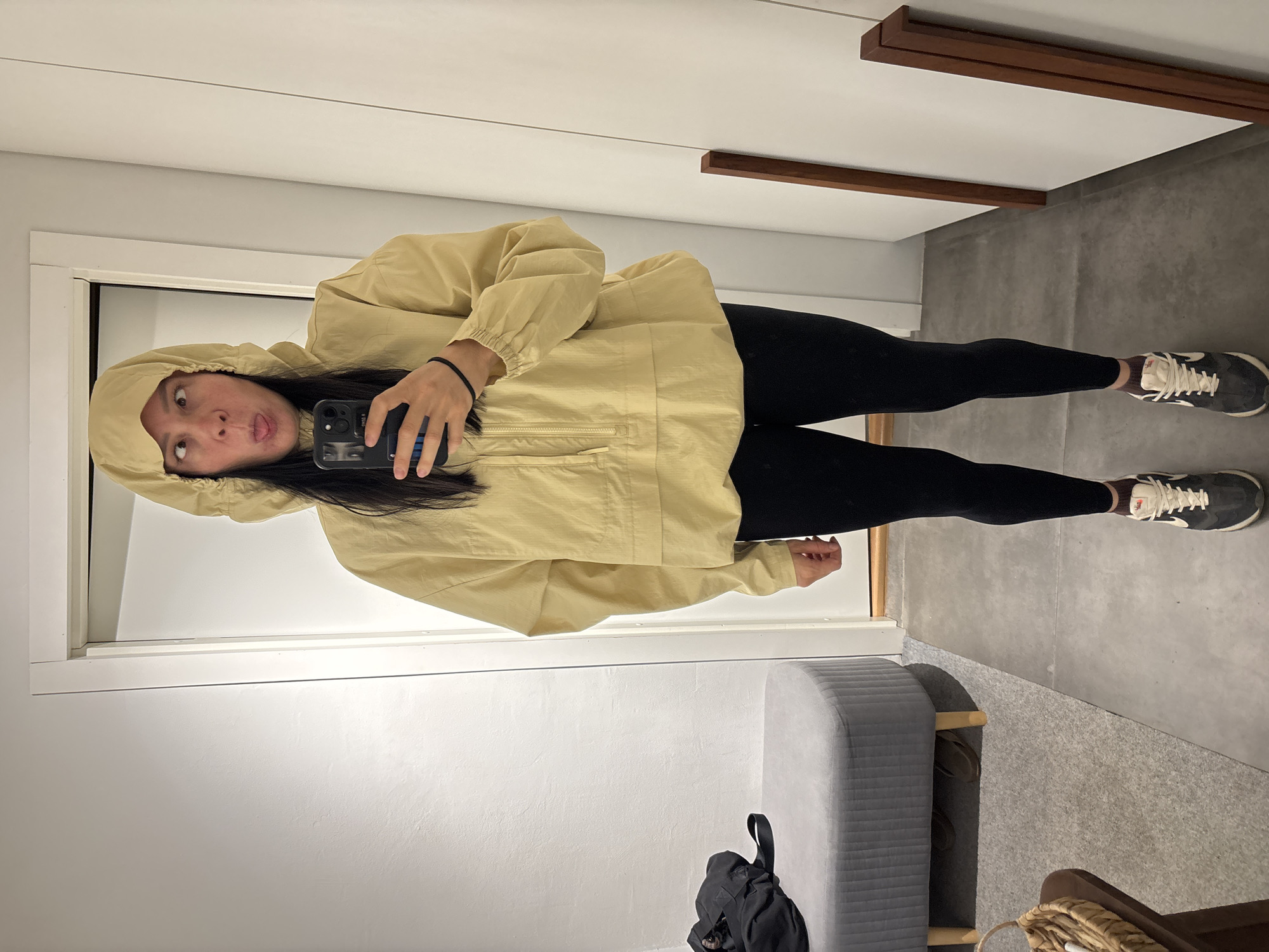 A young woman takes a selfie in a dressing room. She is wearing a light yellow hooded jacket and black leggings. Her hair is long and dark. She is holding a black smartphone in her right hand. The dressing room has a white door and walls, with wooden trim along the top. A gray padded bench sits near a black backpack on the floor. The floor is light gray and there is a glimpse of another room to the right. The photo was likely not taken in Chaikovsky, Russia.