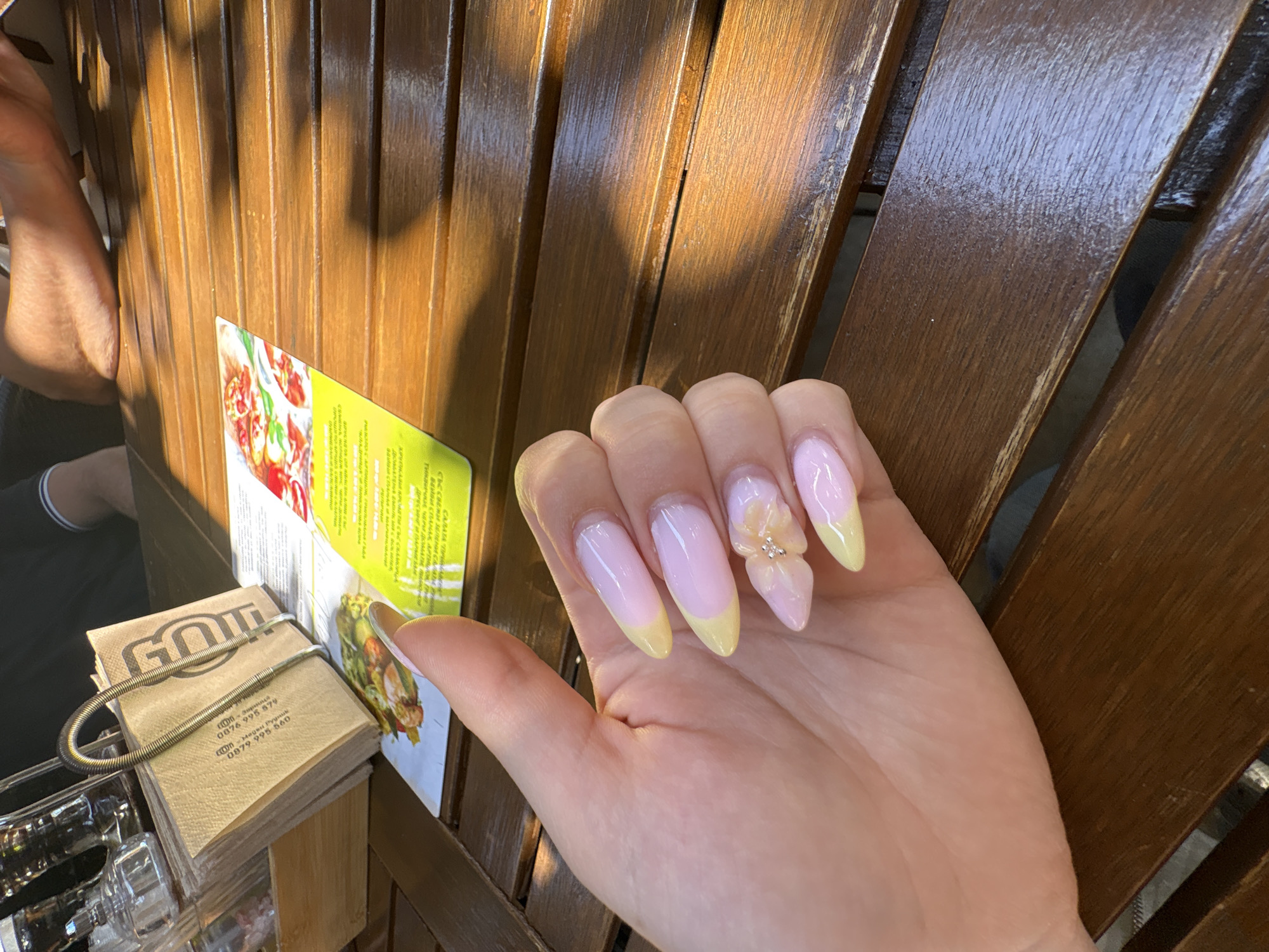 The image features a close-up of a person's hand displaying a fresh manicure against a sun-drenched, wooden background, set in what appears to be a cafe or restaurant in Burgas, Bulgaria. The nails are long, with a light pink base and vibrant yellow tips, styled in a modern French manicure. The ring finger's nail stands out with an intricate 3D flower embellishment adorned with small rhinestones.
Strong, warm sunlight streams across the scene, casting distinct shadows on the wooden slatted surface, suggesting it is a bright, sunny day. In the upper left background, part of another person's arm or leg is partially visible.
To the left of the prominent hand, a stack of tan napkins is held in a spiral metal dispenser. The napkins bear text including "GOL", "000- зарания", "0876 995 579", "сооп Меден Рудник" (a district in Burgas), and "0879 995 560". Next to the napkins, a green-bordered menu or flyer is partially visible, showing appealing food photography, likely of a salad. Visible Bulgarian text on the menu includes "Swaune", "САЛАТА "ПРИВА" (Prival Salad), "СЪС СВЕЖИ ЗЕЛЕНИ САЛА" (With fresh green sala...), "БЕЙБИ СПАНАК, АРТИШОК ВИЯ" (Baby spinach, artichoke...), and "ТИКВИЧКИ ЧАРИ ДОМАТИ" (Zucchini cherry tomatoes). A clear glass container is also visible in the lower left corner. The overall setting implies a relaxed meal or coffee break.