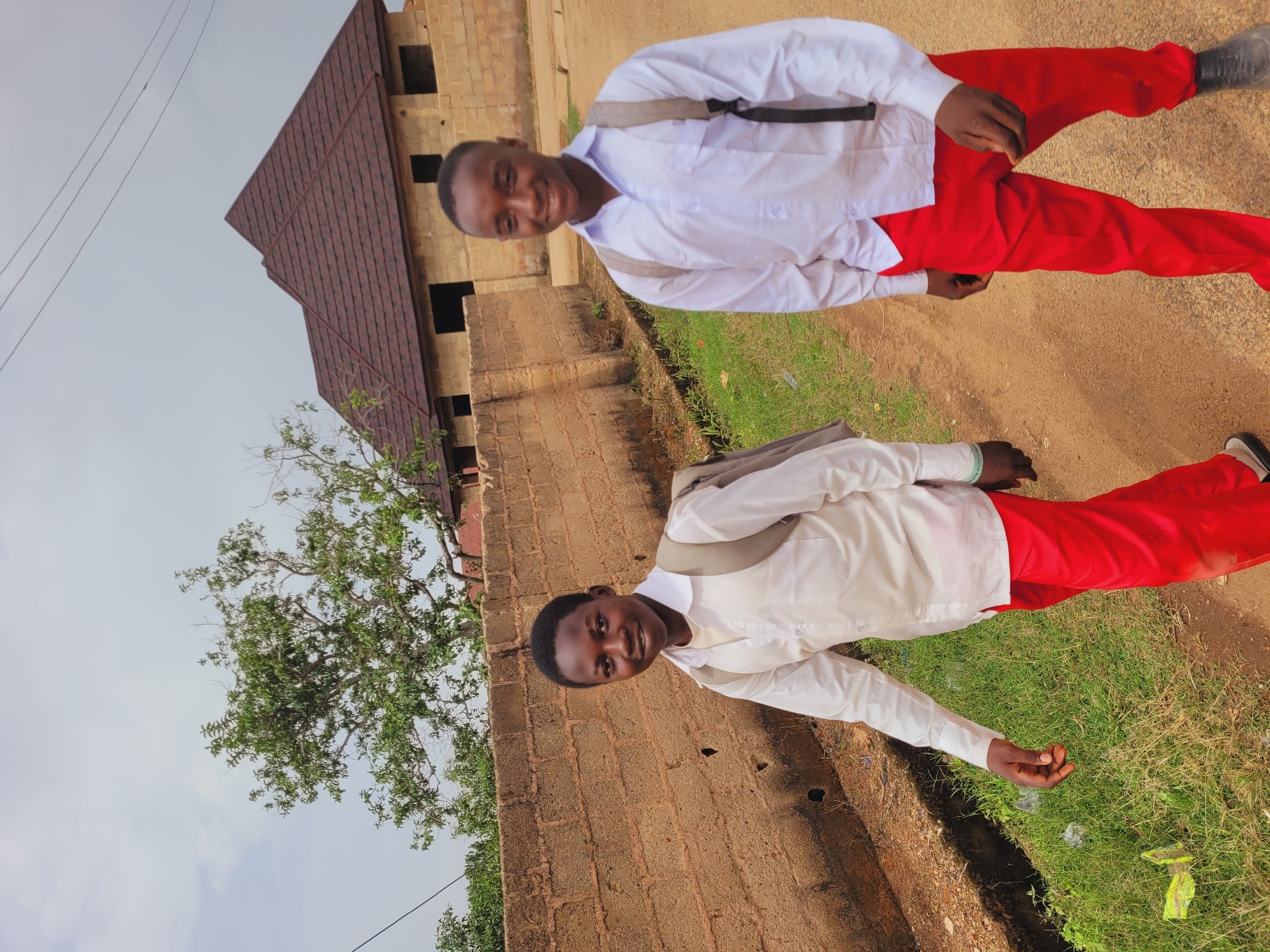 Two young males, appearing to be students based on their uniforms, are walking outdoors. The male in the foreground, on the right, is wearing a white collared shirt and red pants, with a gray backpack. He is looking directly at the camera and smiling. The second male, on the left, is also wearing a white shirt and red pants, with a gray backpack, and is looking slightly away from the camera. They are walking on a dirt path next to a low, rough brick wall. Behind the wall, there is green vegetation. Further in the background, a building with a brown tiled roof and unfinished brickwork is visible, along with a large tree with green foliage. The sky is overcast. This scene is located in Bukuru, Nigeria.