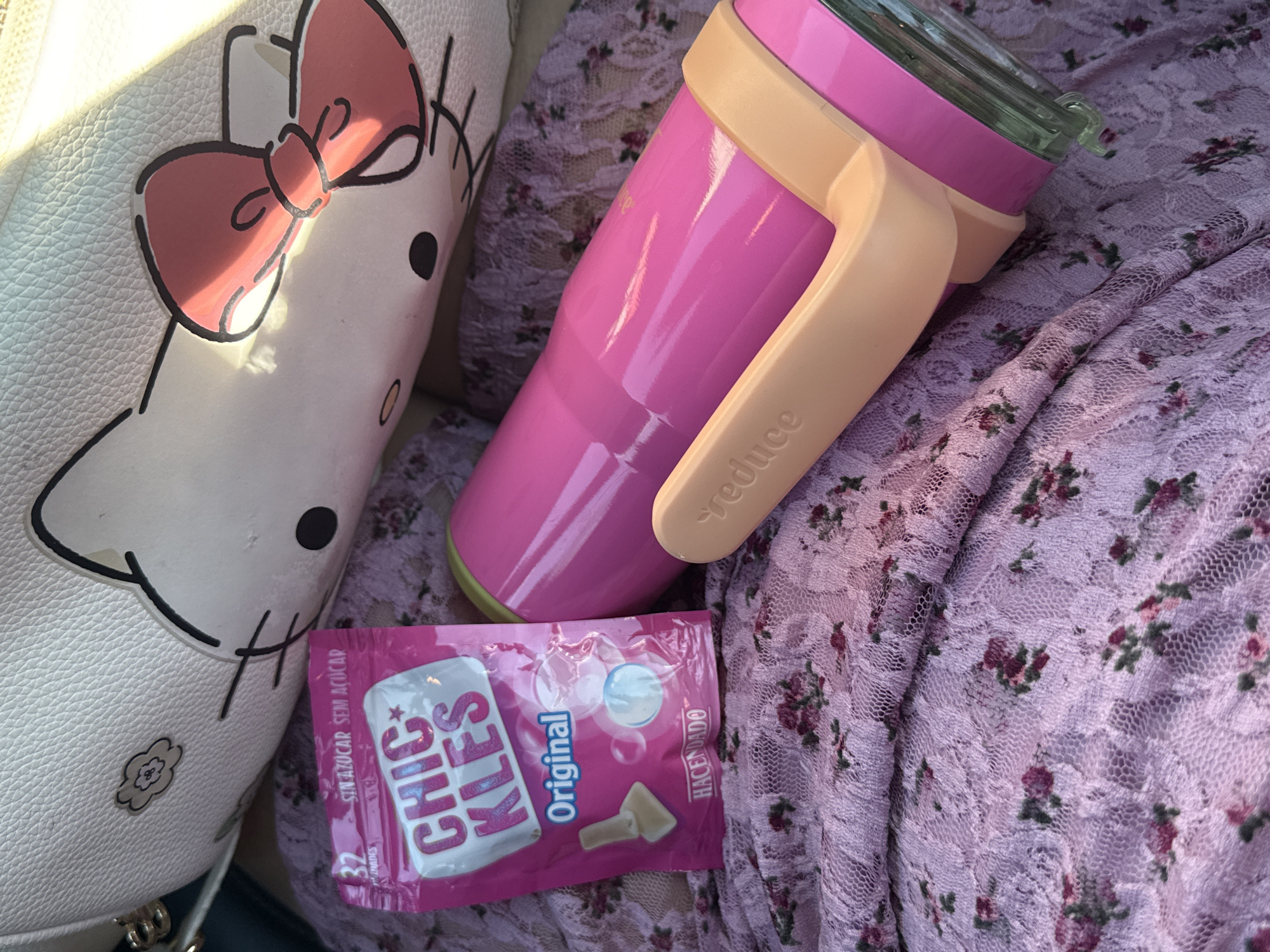 This is a close-up, eye-level, still shot of several items laid out on a purple lace fabric with a floral pattern. In the upper portion of the image is a white handbag with a Hello Kitty design. To the left of the handbag is a pink packet of "Chic Kles Original" gum. To the right of the gum is a bright pink thermal bottle with a beige handle. The bottle has the word "reduce" embossed on the handle. The fabric appears to be a soft, possibly velvet-like material with an intricate lace overlay featuring small dark red and green flowers. The lighting suggests it is daytime, with some bright sunlight casting shadows and highlights on the objects, particularly the Hello Kitty handbag. There are no people or actions depicted in the image. The location is not identifiable as Madrid, Spain.