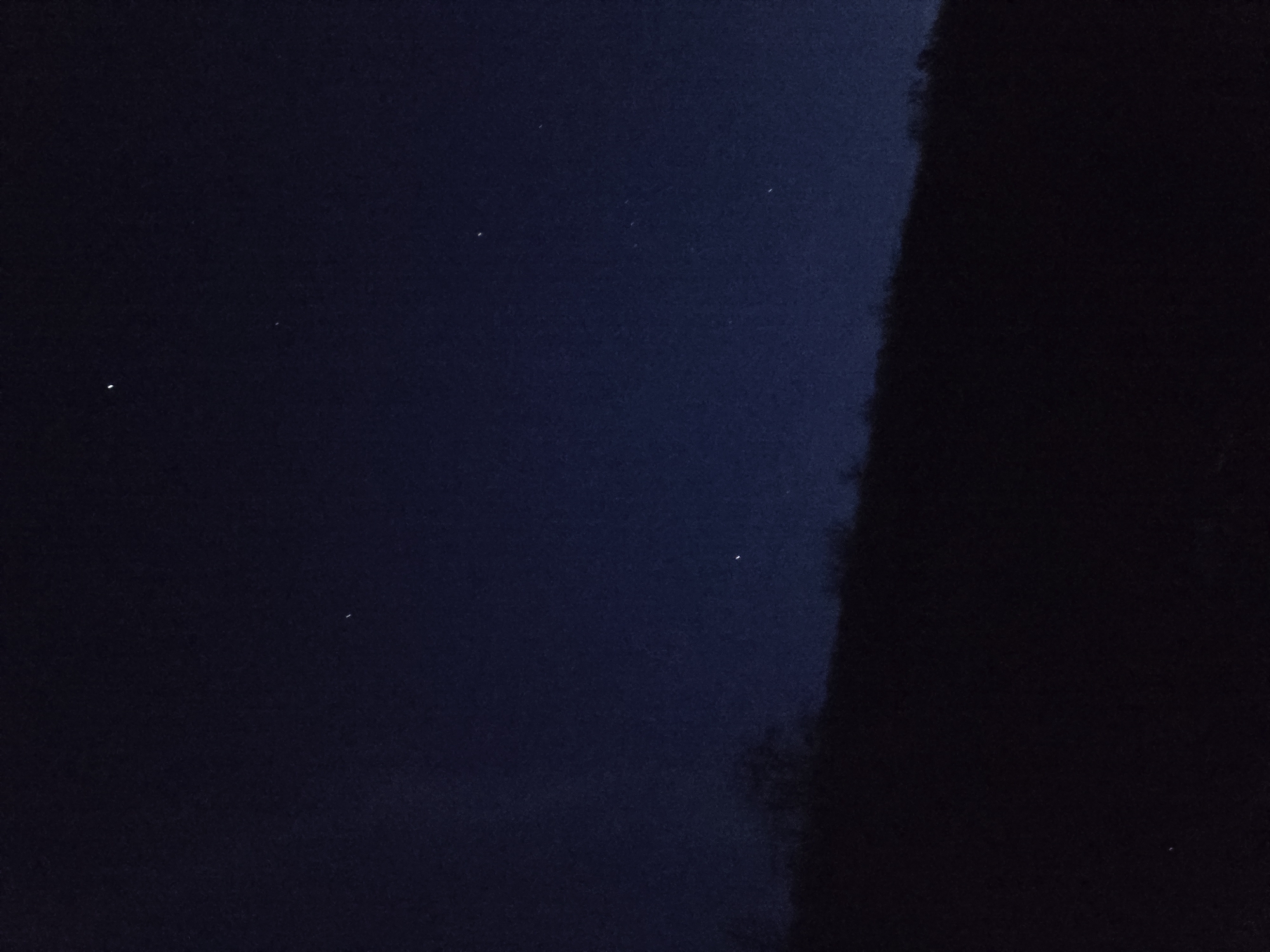 The image depicts a night sky with several visible stars. A dark, indistinct mass occupies the right side of the frame, likely a tree or a building. The sky is a gradient of dark blue, lighter towards the upper left. The stars appear as small, bright dots scattered across the darker background. The location is Unknown, Unknown.