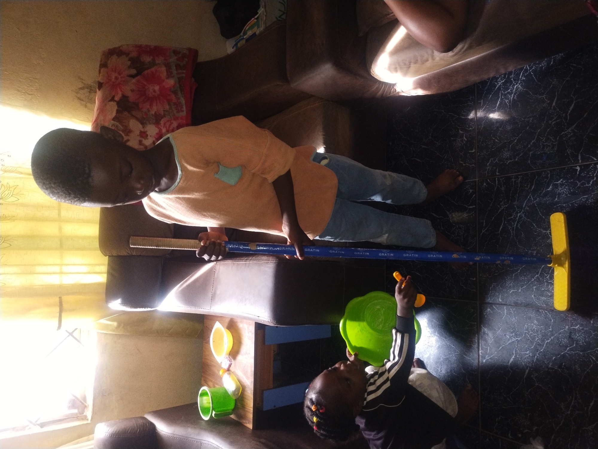 Two young children are in an indoor setting, likely a home. On the left, a toddler with braided hair is crouched on a dark, tiled floor, reaching into a bright green bowl. They are wearing a black and white tracksuit. To the right, a slightly older child stands holding a blue and yellow broom. They are wearing an orange shirt and blue jeans. In the background, two brown leather couches are visible, one with a patterned fabric draped over its back. A small wooden table with a green cup and a yellow bowl sits between the toddler and the couches. Sunlight streams in from a window behind the couches, casting shadows on the floor. The overall impression is of a domestic scene, possibly during playtime or a chore.