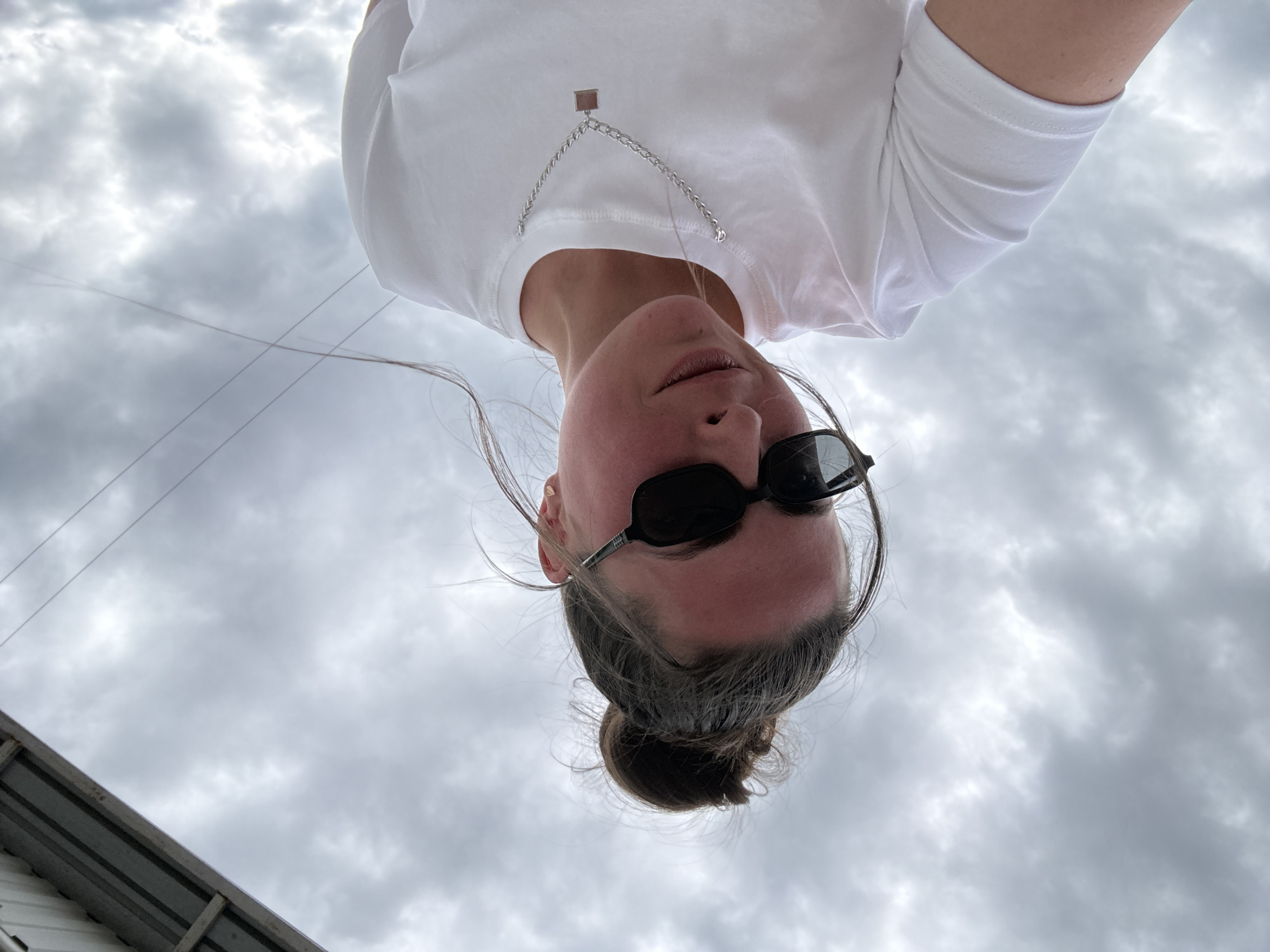 A low-angle medium shot shows a young adult with their hair in a messy bun, wearing sunglasses and a white t-shirt. The subject is looking down slightly and has a subtle smile. A silver chain with a small square pendant hangs around their neck. The background is a cloudy sky with a sliver of a structure overhead. The location is unknown.