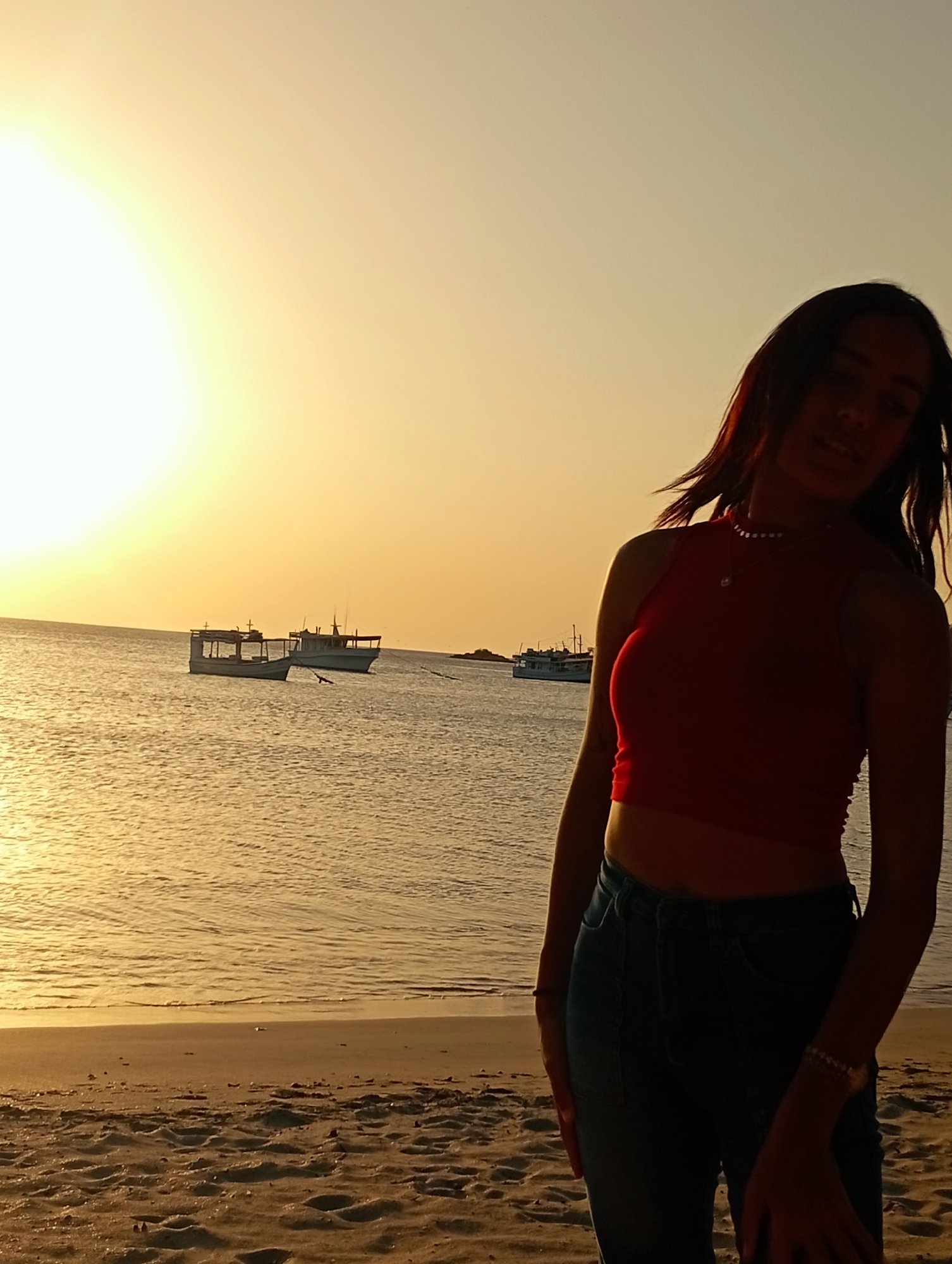 The image shows a woman standing on a sandy beach. She is wearing a red tank top and dark jeans. The location is Juan Griego, Venezuela, indicated by the beach environment and the presence of fishing boats. The sun is setting, casting a warm glow over the scene, and the sky is a gradient of yellow and orange. In the background, several boats are visible on the water. The beach appears to be relatively empty, with just the woman present.