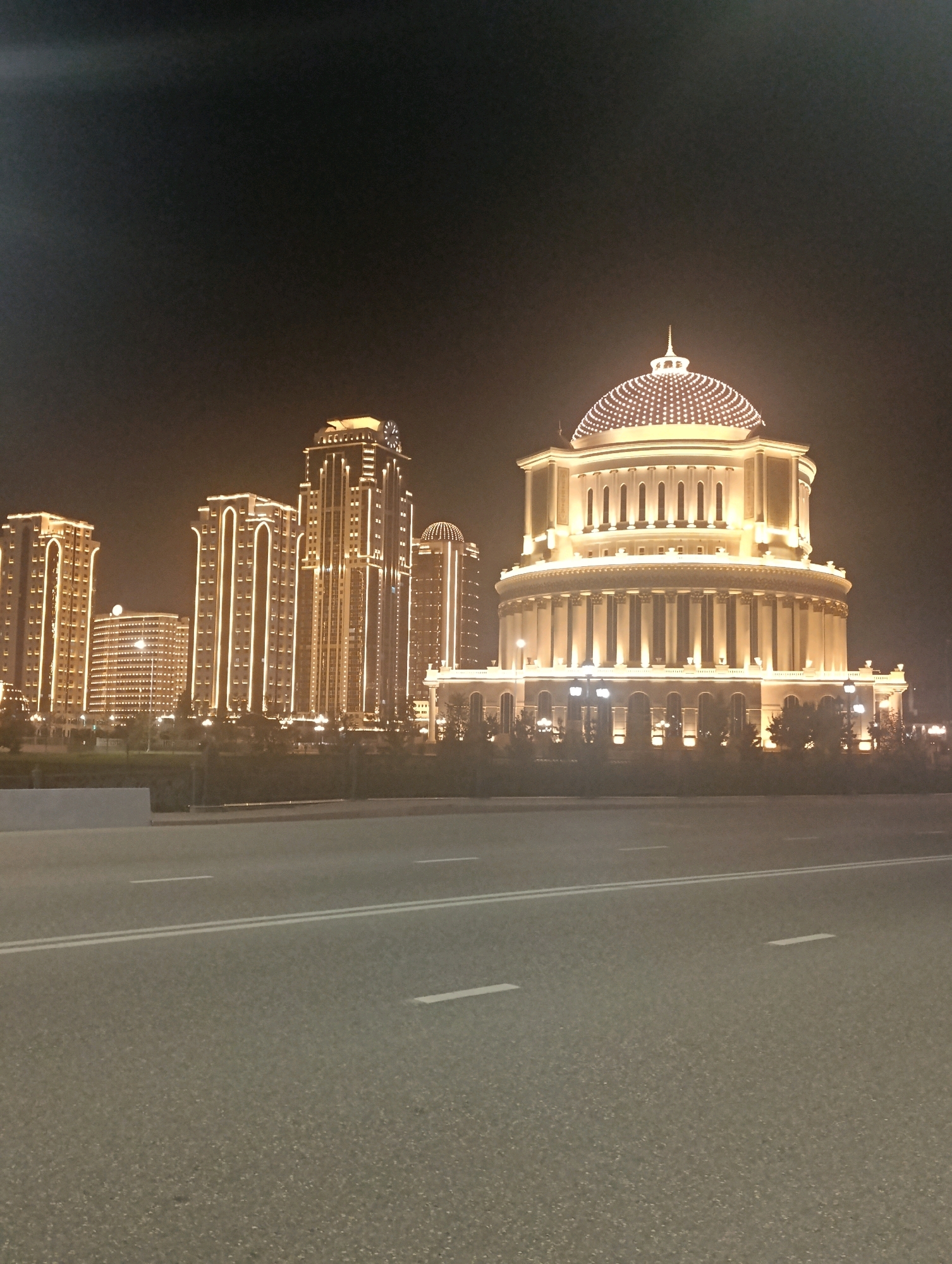 The image depicts a night scene in a city, likely in a developing country or an area undergoing rapid modernization. The foreground is dominated by a dark asphalt road with white lane markings. In the background, illuminated by warm, yellow lights, is a collection of modern high-rise buildings and a prominent, domed structure. The high-rise buildings are tall and slender, with vertical lines of lights tracing their edges and outlining their architectural features. One of the buildings has a clock face at its summit. The domed structure is grand and classical in design, featuring a row of large columns encircling its base and a patterned, illuminated dome topped with a spire. The overall impression is one of urban development and illuminated nighttime architecture.