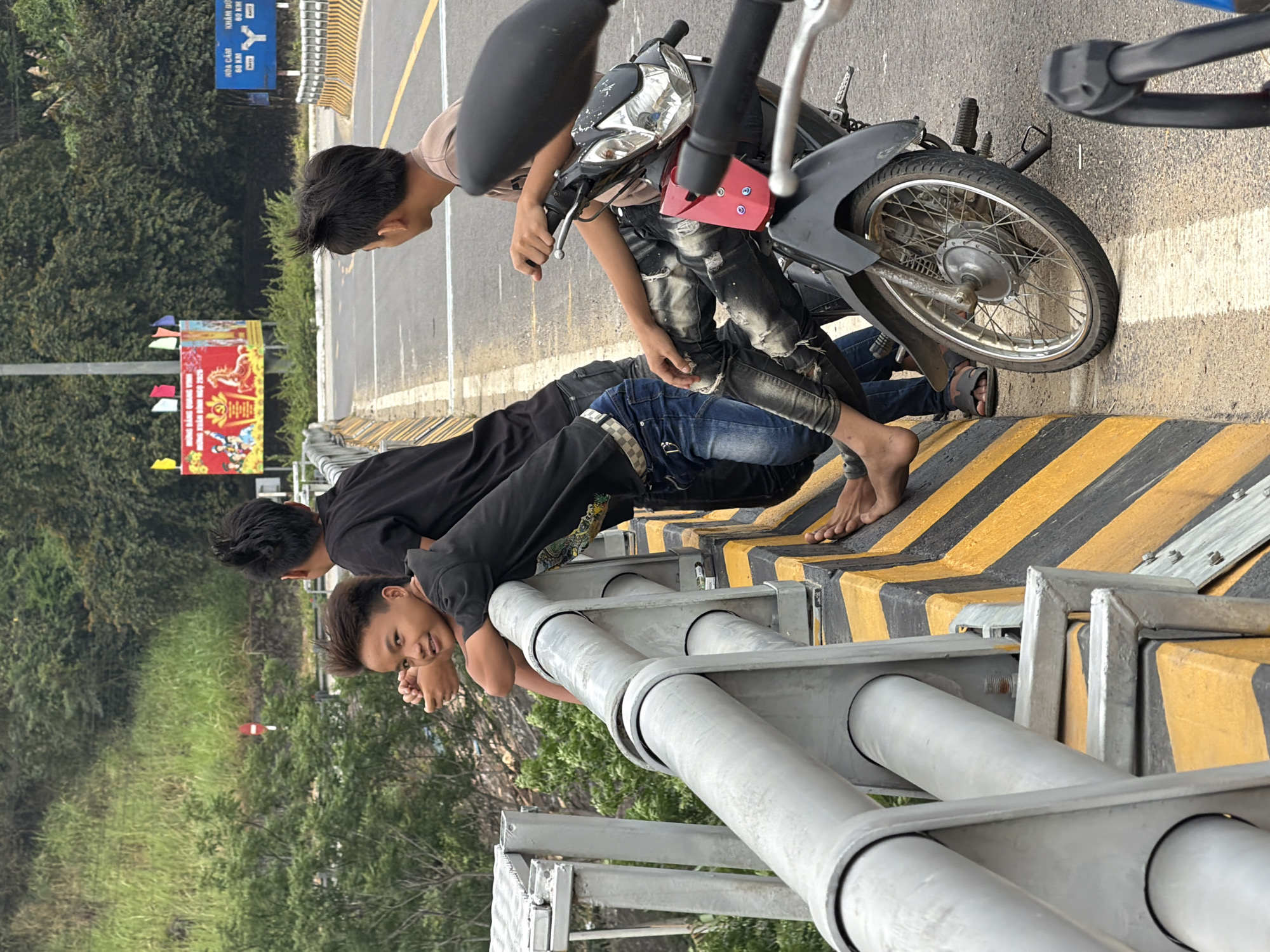 Three young men are on a bridge in Đà Nẵng, Vietnam. One sits on a parked motorcycle, another is leaning against a railing of the bridge, and the third is behind. All are partially barefoot and leaning over the edge of the bridge. The environment includes foliage, guardrails, and signage.