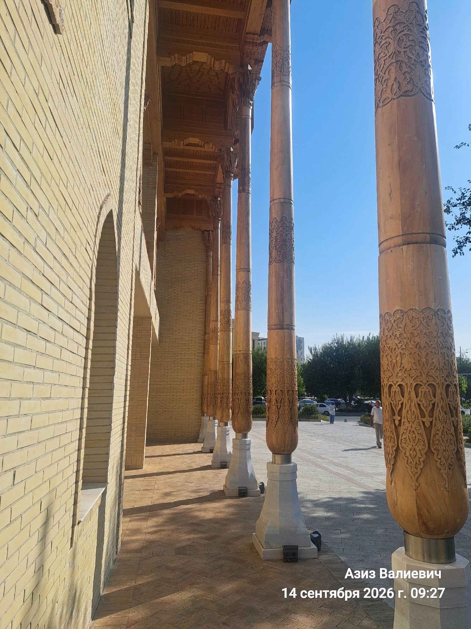 An outdoor shot captures a row of ornately carved wooden columns supporting a covered walkway. The columns are light brown, cylindrical, and feature intricate patterns carved into their surfaces. They stand on white, multi-tiered bases. The walkway is paved with tan bricks laid in a herringbone pattern. To the left, a wall constructed of pale yellow bricks runs parallel to the columns, featuring arched openings. Sunlight casts long shadows across the walkway and the brick wall. In the background, trees and buildings are visible, and two individuals are walking away from the camera on a paved area. The sky is clear and blue. The image is overlaid with text in Cyrillic script which reads: "Азиз Валиевич 14 сентября 2026 г. 09:27". This text indicates the date and time the photo was taken. The location is the Minor Mosque (Minor Masjidi) in Tashkent, Uzbekistan.