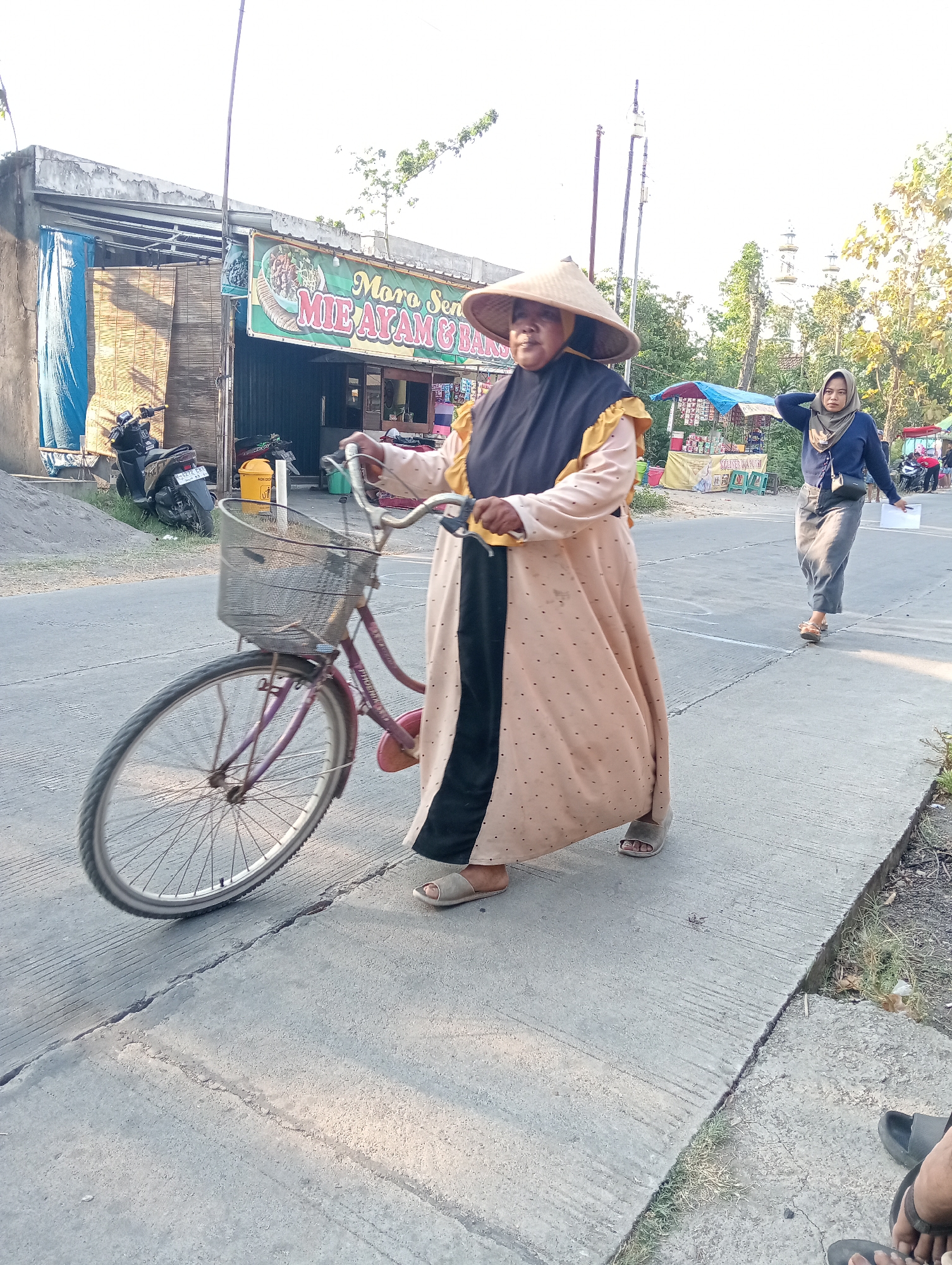 A middle-aged woman, wearing a conical hat and a long, light-colored dress with dark trim, walks a bicycle on a paved road in Tanjunganyar, Indonesia. The bicycle has a wire basket in front, which appears empty. Behind her, further down the road, another woman wearing a hijab and a long, dark blue top and grey skirt walks away from the camera, with her right hand touching her head. A building with a sign reading "Moro Sen MIE AYAM & BAKSO" is visible to the left of the frame. Various stalls and parked motorcycles are also present in the background. The scene is illuminated by natural sunlight.