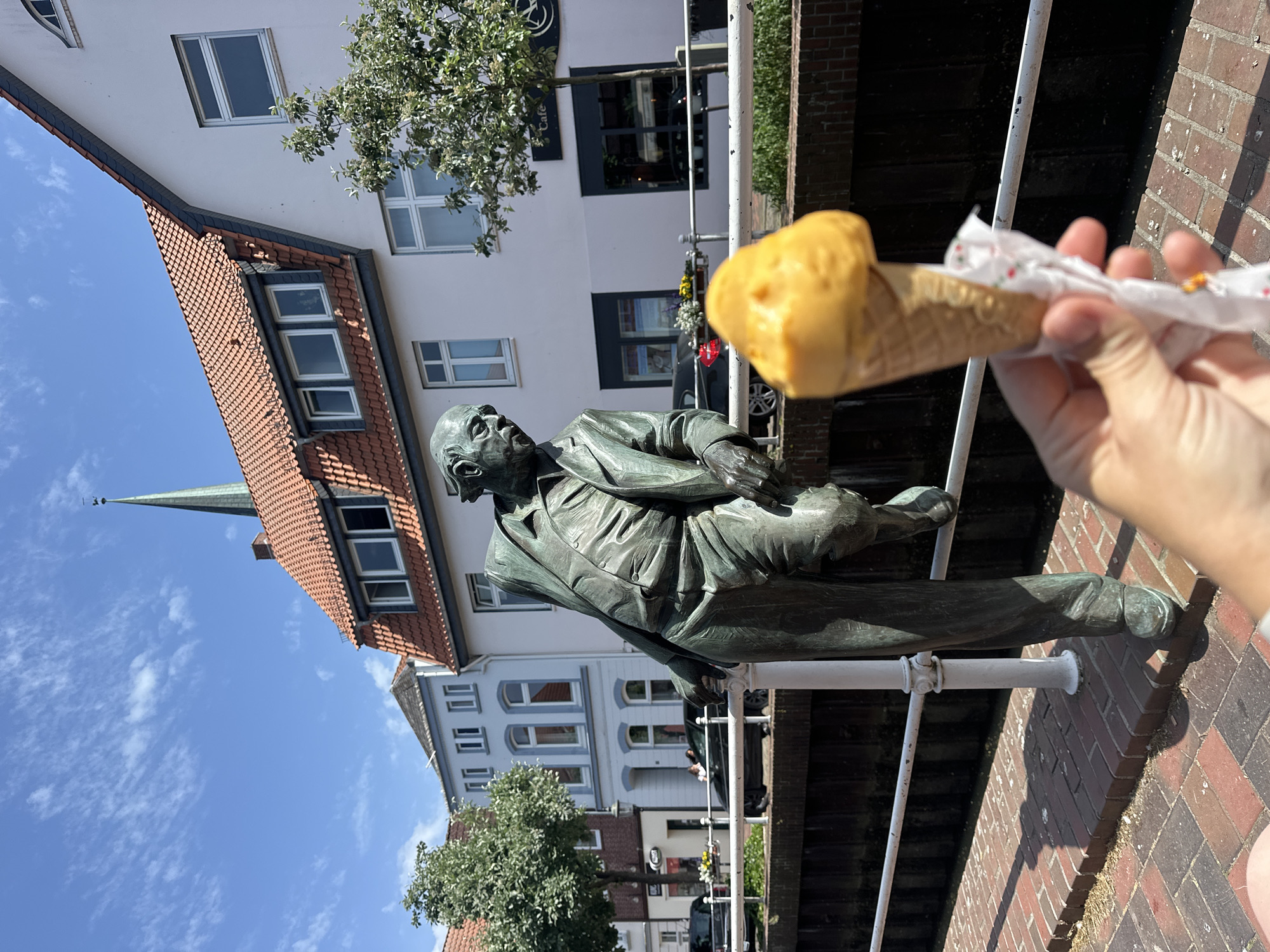 A statue of Wilhelm Külchen is positioned near the canal and buildings in Buxtehude, Germany. In the foreground, a hand holds a wrapped ice cream cone. The photo is taken on a sunny day, as evidenced by the shadows and blue sky.