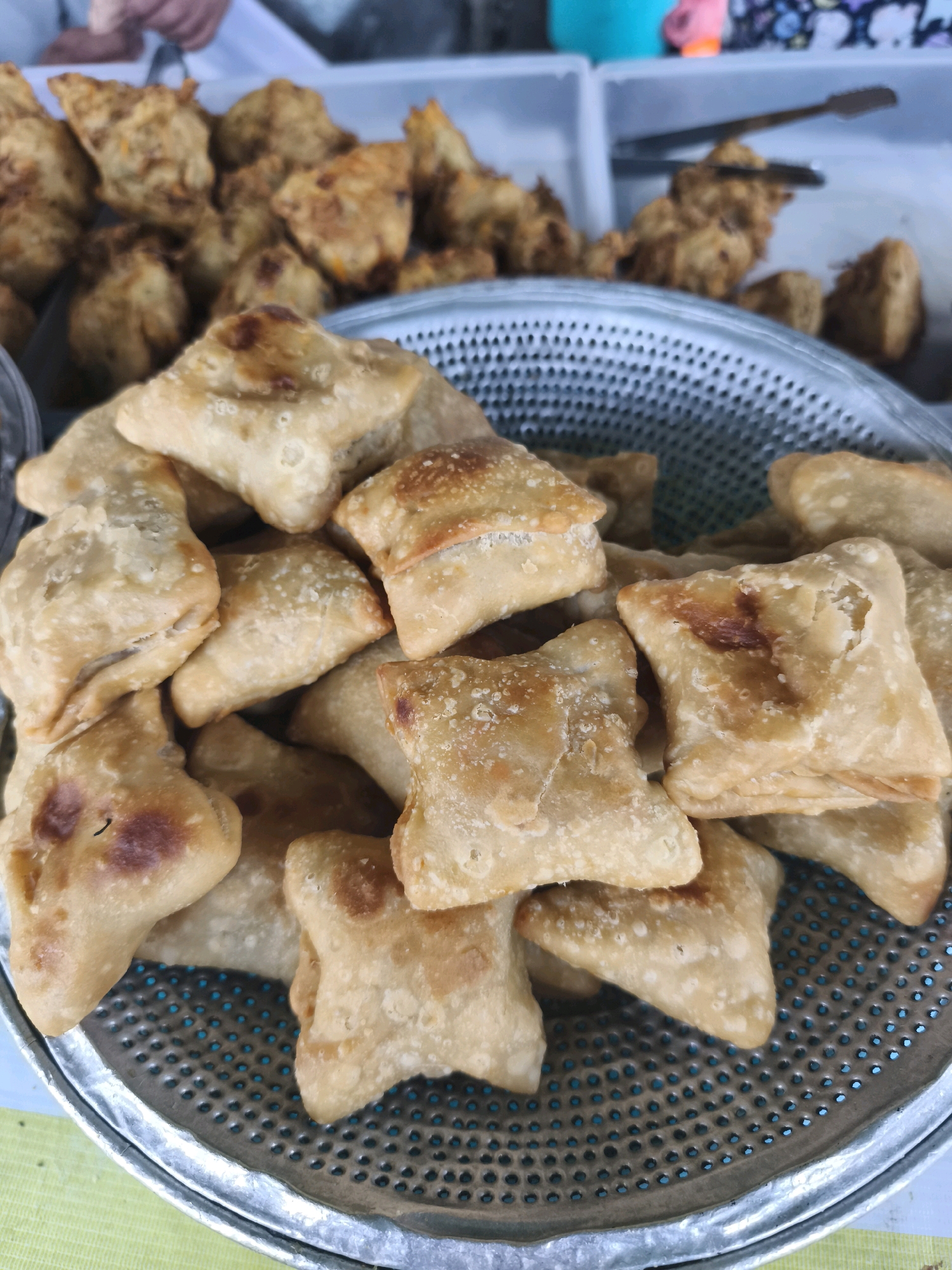A close-up, eye-level shot shows a metal colander filled with fried pastries. The pastries are golden brown and appear to be of various shapes and sizes, with some triangular and others more square-like. They have a flaky texture and some show darker, crisped edges. In the background, out of focus, are more pastries in white containers, some being handled with tongs by an unseen person. The overall scene suggests a food stall or market in Palembang, Indonesia, likely selling these freshly fried snacks. The lighting is natural and diffused, highlighting the appetizing appearance of the food.