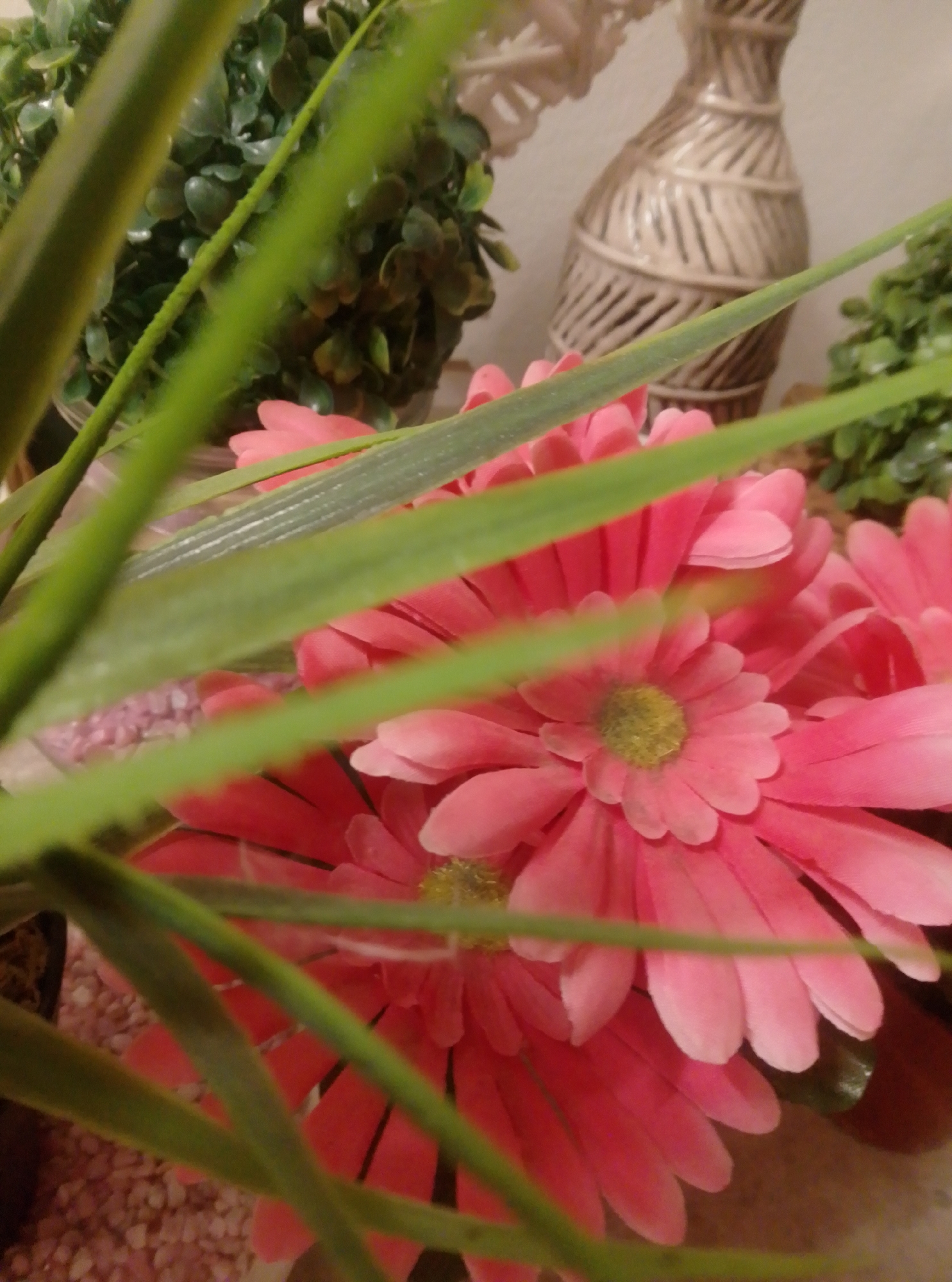The image shows a close-up view of several artificial pink gerbera daisies with green, grass-like foliage in the foreground. In the background, there is a decorative ceramic vase with a textured, woven pattern. Behind the vase, there are two artificial green plants. The setting appears to be indoors, possibly a decorative display in a home or shop. There are no people or animals visible in the image. The image does not provide any specific information that could be used to determine its location in the city of Wedel, Germany.