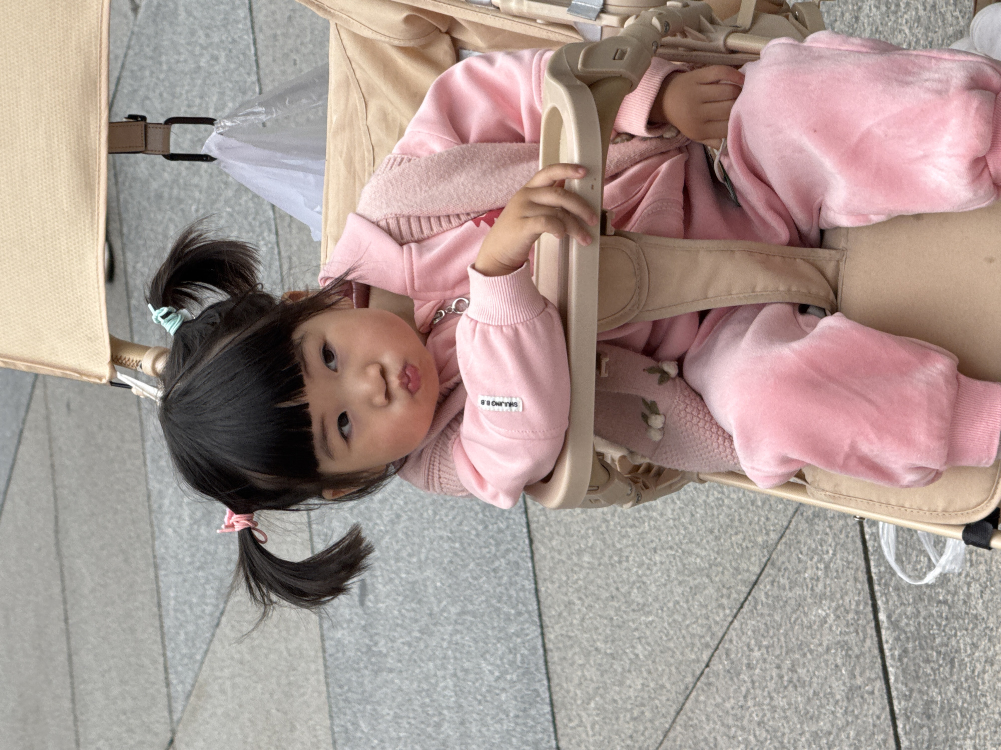 A toddler with black hair styled in pigtails with colored hair ties sits in a beige stroller. The child is wearing a pink outfit and faces the camera with a slight pout. The stroller has a beige harness. The backdrop is a grey paved surface, presumably a sidewalk. The location is potentially Jinjiang District, China.