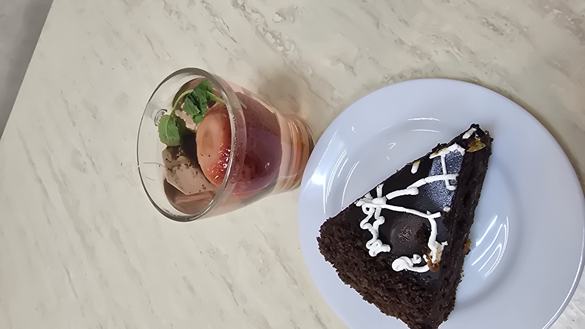 A slice of chocolate cake sits on a white plate, next to a clear glass filled with a pinkish liquid, pieces of chocolate, and strawberries. The cake is dark brown with a textured outer layer and is decorated with white icing. The glass contains what appears to be ice cream, strawberries, and mint leaves, with a light pink liquid base. Both items are placed on a light-colored, marbled surface. The image does not provide any information or context to determine the location in the city of Kazan, Russia.