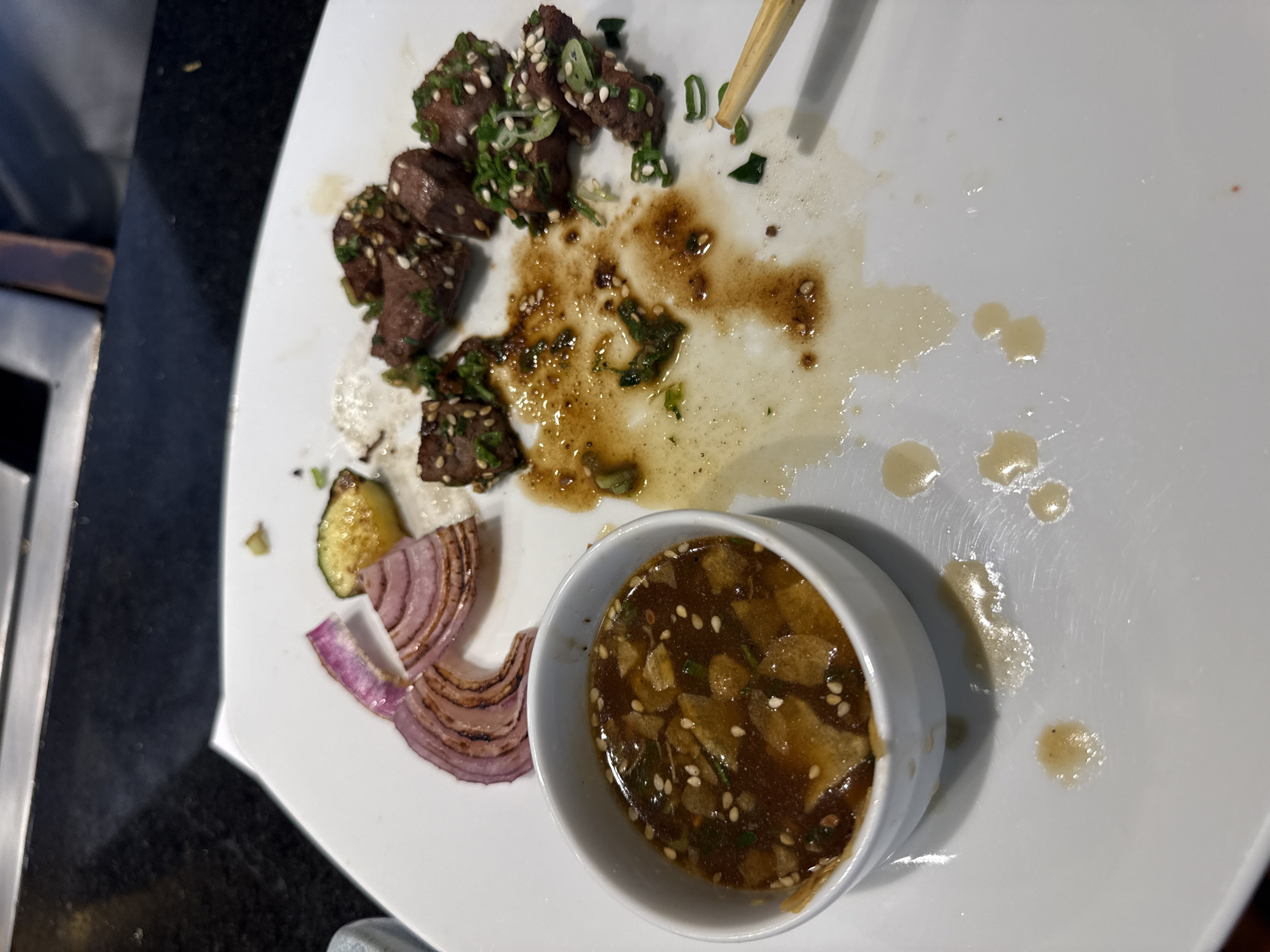 A partially eaten meal sits on a white, square plate. Leftovers include cubed beef, sesame seeds, green onions, grilled red onion slices, and a small bowl of sauce containing visible sesame seeds and scallions. The plate also shows residual sauce and scattered food remnants. A pair of chopsticks rest on the plate near the beef. The setting appears to be indoors, likely in a restaurant. There is no verifiable information about the location being Puebla, Mexico, within this image.
