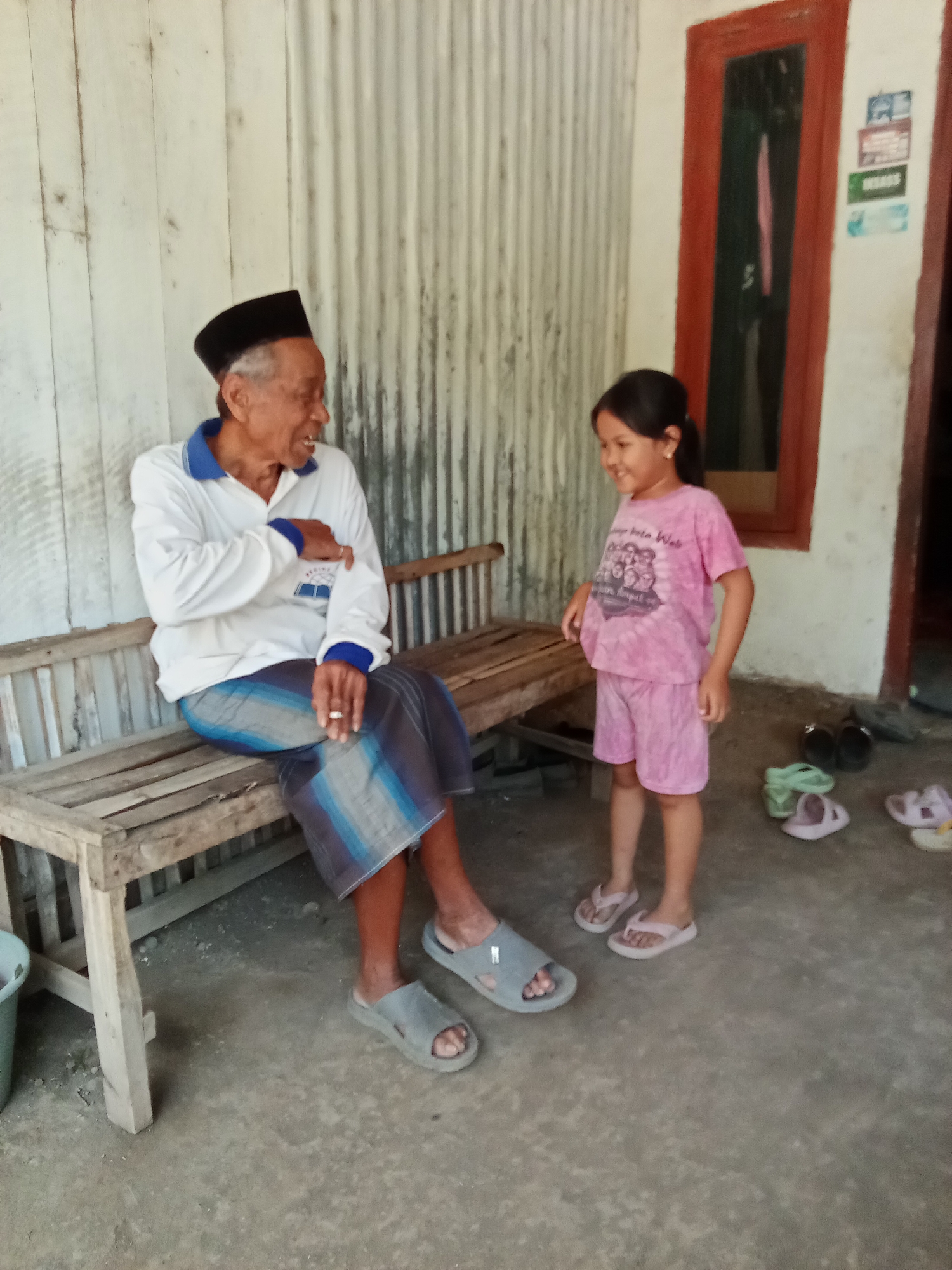 An elderly man wearing a black peci and a white long-sleeved shirt with blue trim, and a patterned sarong, sits on a wooden bench. He is holding a cigarette in his right hand. A young girl in a pink t-shirt and shorts stands facing him, smiling. Behind them is a corrugated metal wall and a building with a red-framed window. The ground is concrete. Several pairs of sandals are scattered on the ground in front of the girl. The scene appears to be outdoors, likely on a porch or patio. The location is identified as Trebungan, Indonesia.