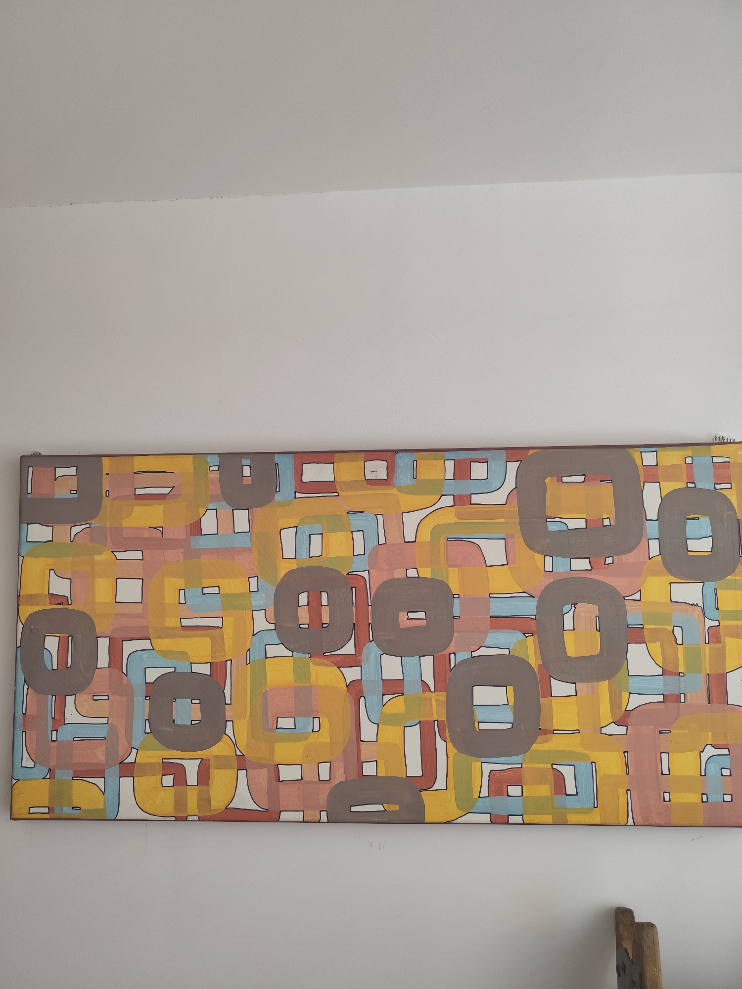 The image shows a long, horizontal abstract painting mounted on a white wall. The painting is characterized by a repeating pattern of rounded square shapes and interlocking lines in various colors, including yellow, pink, blue, brown, and white. The colors are applied in a somewhat flat, opaque manner, with black outlines defining the shapes. The painting is framed by a thin, dark border. The wall appears to be painted a matte white. In the lower right corner of the image, a portion of a wooden object, possibly part of a chair or stand, is visible. The lighting suggests an indoor environment, with soft, ambient light. There are no people or discernible actions taking place in the image. The overall impression is of a decorative art piece in a simple setting.