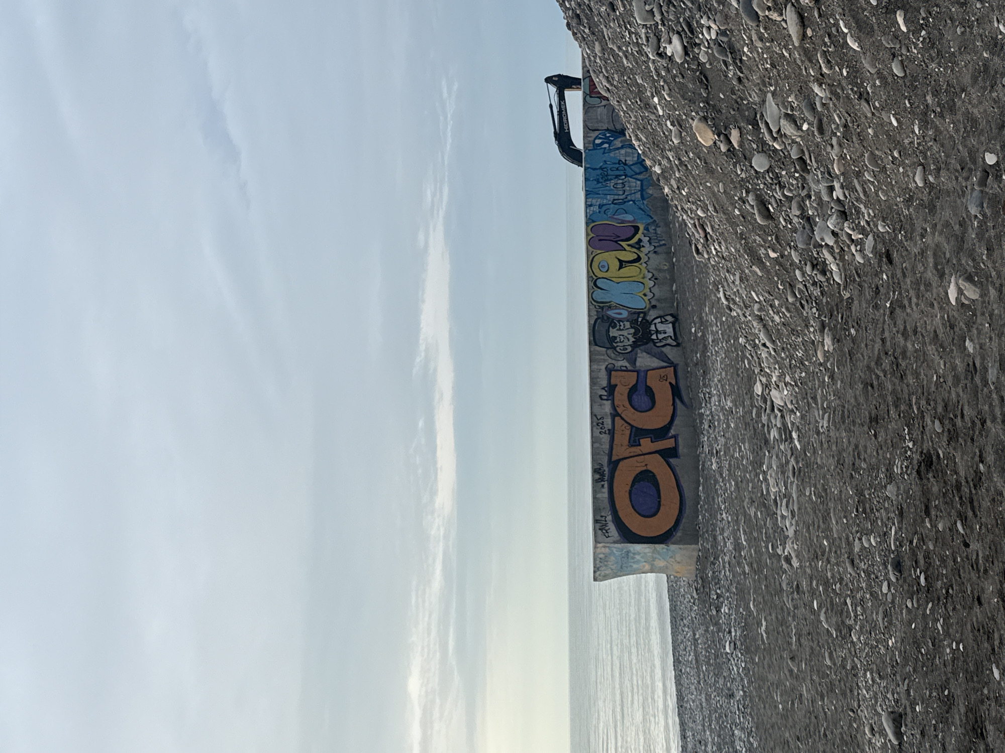The image depicts a concrete structure covered in graffiti, located near a rocky embankment and the sea. The sky is visible in the background. The structure bears graffiti in multiple colors, including blue, yellow, purple, and orange. The graffiti includes stylized letters and cartoonish faces. Above the graffiti, there is some construction equipment with the name "HIDROMEK" written on it. The rocky embankment consists of small, dark-colored pebbles. The sea appears calm and stretches to the horizon. The location is likely in or near Batumi, Georgia, given the presence of concrete sea defenses along the coastline.