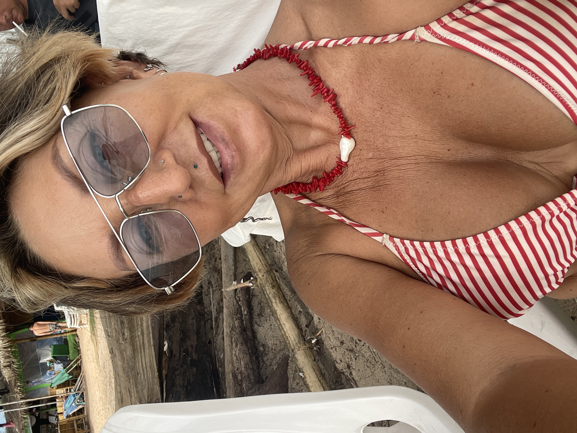 A woman is captured in a close-up selfie on a sandy beach, likely in Choeng Thale, Thailand, as indicated by the background details. She is wearing a red and white striped bikini top and a red coral necklace with a white pendant. Her blonde hair is styled short, and she wears large silver-rimmed sunglasses. Visible piercings include one on her septum and others on her ears. The background reveals a section of a beach hut with a thatched roof, wooden structures, and beach chairs, suggesting a tropical resort or public beach area. The sand and driftwood in the foreground indicate a natural beach environment. The lighting suggests it is daytime, possibly mid-day, given the brightness and the shadows.
