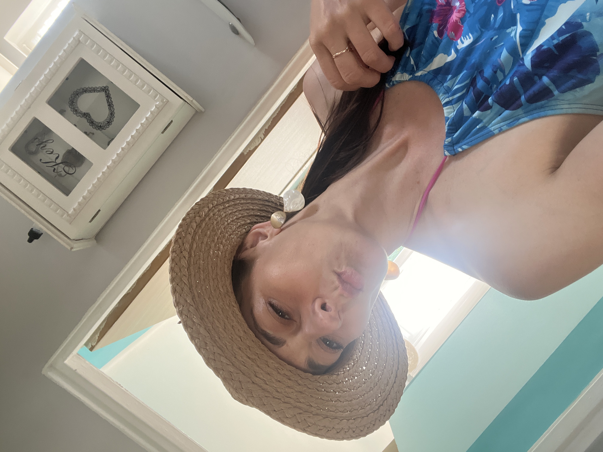 A low-angle, close-up, selfie-style shot shows a woman wearing a straw hat and a blue floral swimsuit. She is puckering her lips as if for a kiss. The woman has long, dark hair that is pulled back and falls over her shoulder. She is wearing large, white earrings and a delicate gold ring on her right hand. Her left arm is visible, resting on her chest. In the background, a light blue wall with white stripes is visible on the left, and a white cabinet with two glass-fronted compartments is on the right. One compartment has the word "Keys" written on it, and the other contains a heart-shaped ornament. The overall lighting suggests a bright, possibly indoor, environment.