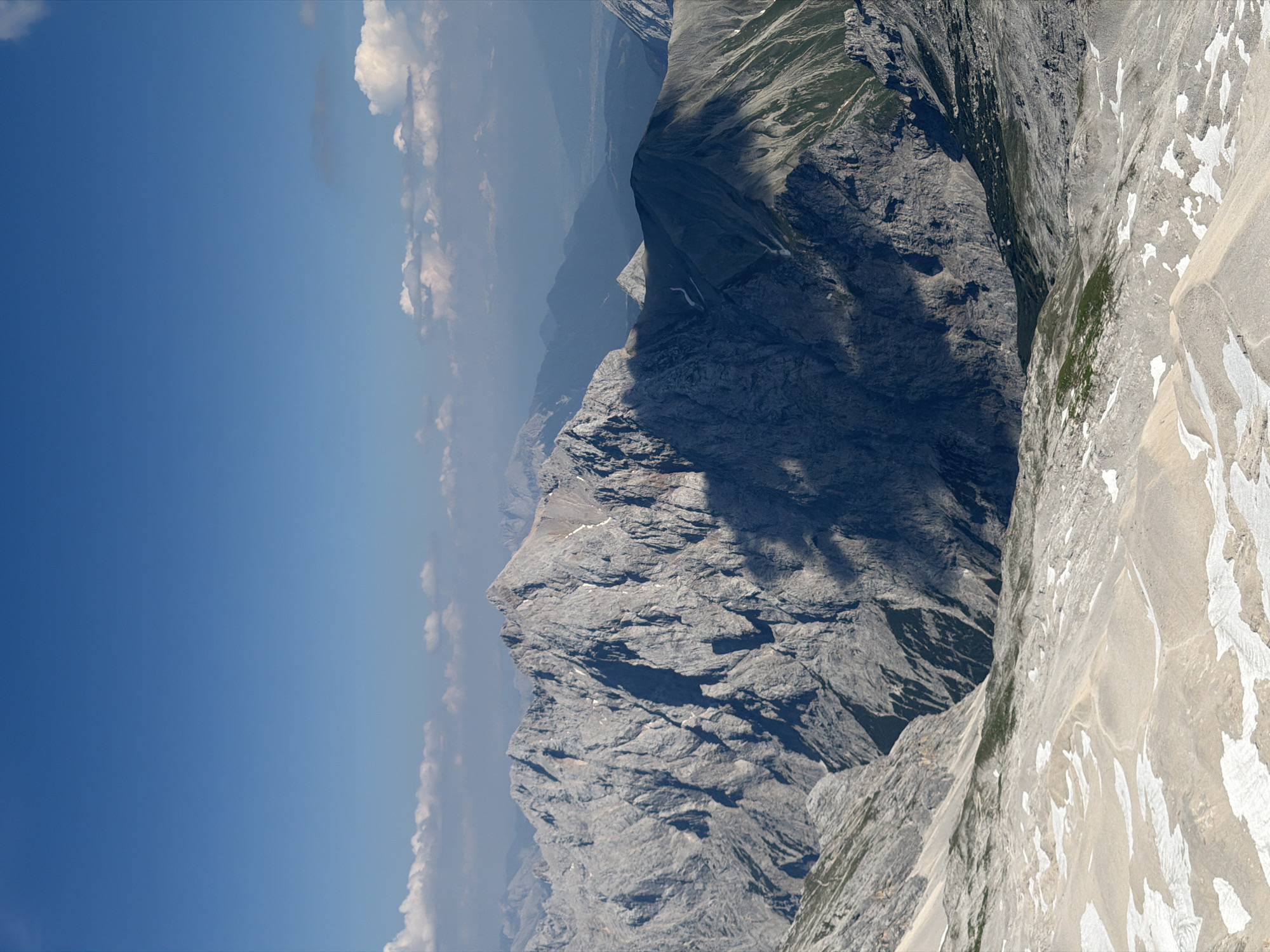 This media file captures a sweeping, high-angle view of a majestic mountain range under a clear blue sky. The scene is dominated by rugged, grey rock formations, carved by time and weather into steep slopes and dramatic peaks. Patches of snow are visible on some of the lighter-colored rock faces, indicating a high altitude.
The landscape transitions from the stark, rocky terrain of the foreground to softer, greener slopes further in the distance, interspersed with dark shadows cast by the sun. The sky is a vibrant blue, with a few wispy white clouds drifting on the horizon, suggesting fair weather. The light implies it is daytime, likely mid-morning or early afternoon, given the strong shadows.
There are no people or man-made objects visible, emphasizing the raw, natural beauty of the alpine environment. The overall impression is one of vastness, grandeur, and the serene power of nature. The location context points to Garmisch-Partenkirchen, Germany, a region renowned for its stunning mountain scenery. The image conveys a sense of awe and tranquility, inviting the viewer to contemplate the scale and beauty of the natural world.