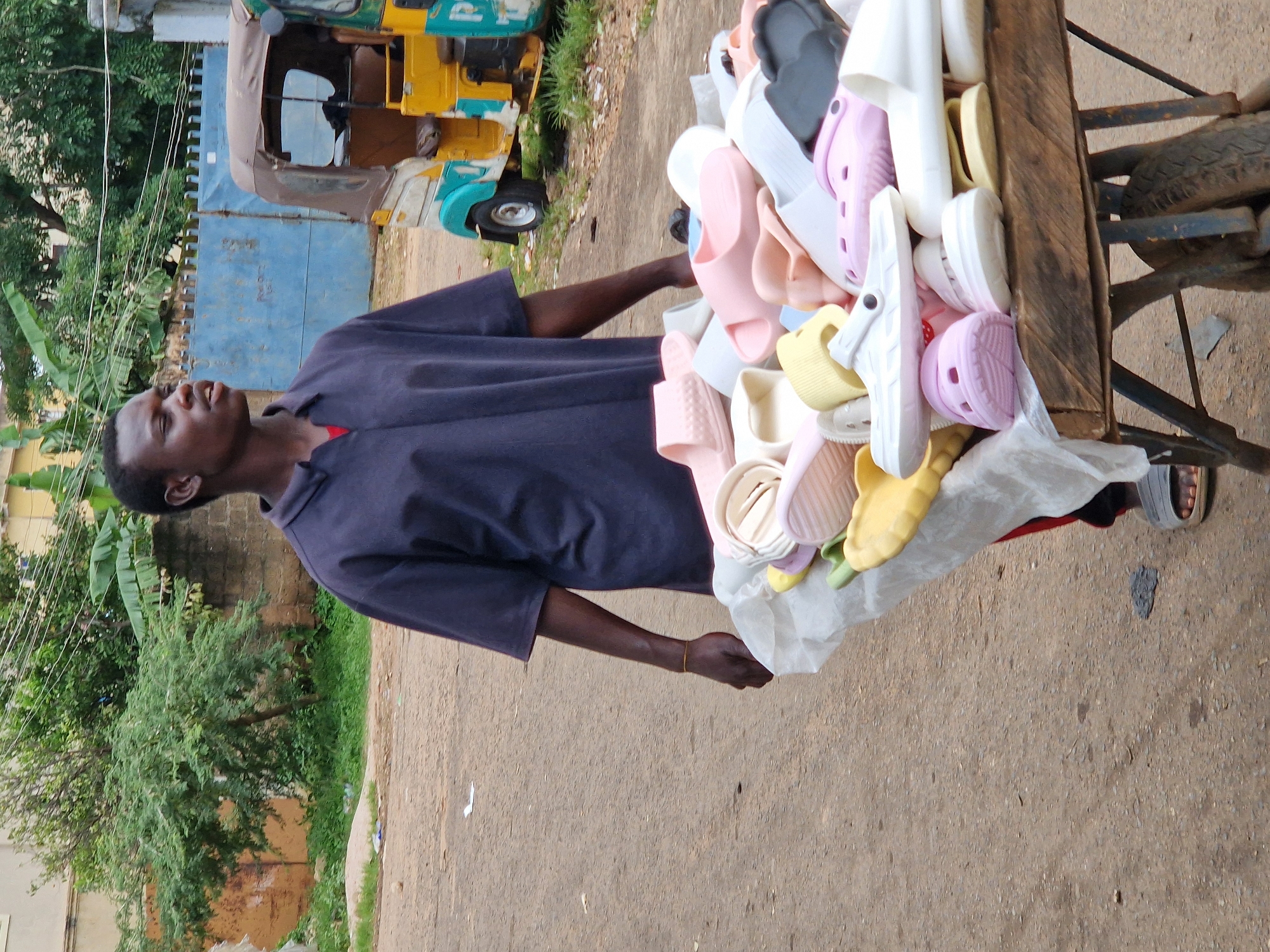 A young Black man is pushing a wheelbarrow full of colorful plastic flip-flops and slides down a dusty, unpaved road. The man is wearing a dark blue collared shirt and dark pants. The wheelbarrow is made of wood and metal and has two tires. The flip-flops are in various colors, including pink, yellow, white, and black. In the background, there is a blue metal gate, a yellow and blue motorized rickshaw, and lush green trees. The scene appears to be in Jos, Nigeria, a city known for its hilly landscape and cooler climate.