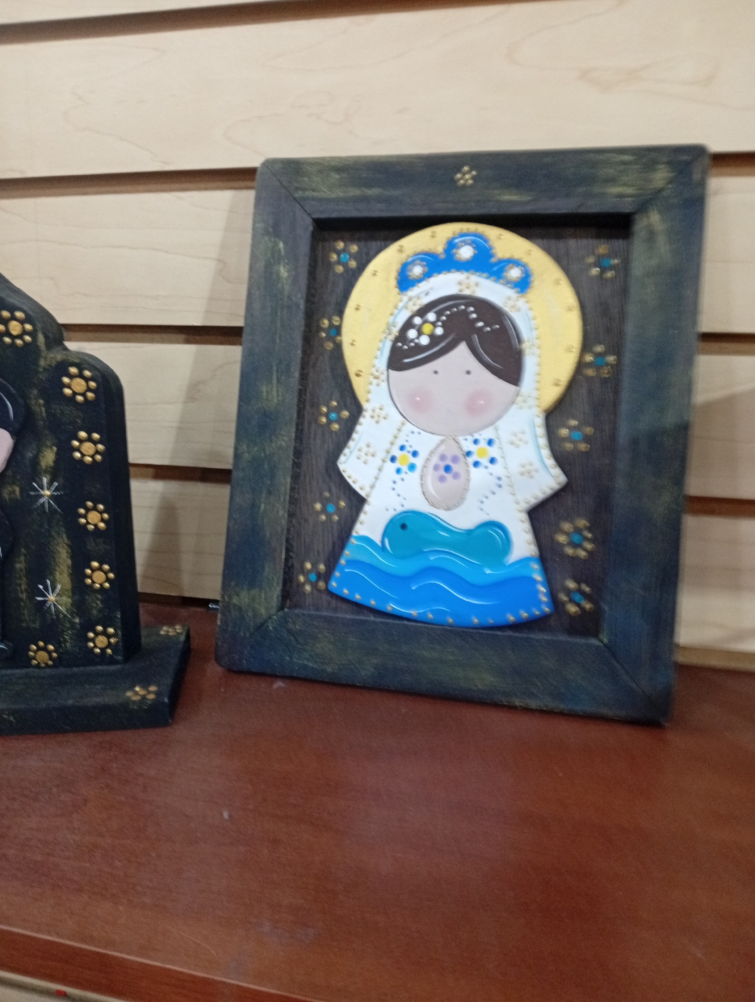 The image shows two religious folk art figures displayed on a wooden surface. The primary subject, to the right, is a framed painting or cutout of a stylized depiction of the Virgin Mary. She is shown with a golden halo, a blue and white robe adorned with small yellow and blue dots, and a light pink face with rosy cheeks. She holds a light purple oval shape in her chest area, and a blue wavy form, possibly representing water or a serpent, is depicted below her. The frame is dark, possibly distressed wood, with a textured finish.
To the left of the Virgin Mary figure is another, taller folk art piece, also likely religious in nature. It is dark colored with gold floral or dot embellishments and appears to have a more angular, sculptural form compared to the framed piece. It is partially visible, but seems to be a standing figure or object.
The background consists of horizontal wooden planks, typical of a wall or partition. The figures are placed on a polished wooden surface, likely a shelf or tabletop, which reflects some light. The scene appears to be indoors, possibly in a shop or home setting. There is no indication of the specific location within Juan Griego, Venezuela, but the style of folk art might be suggestive of the region.