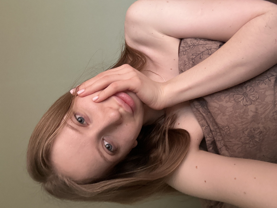 A female individual, likely in her early to mid-twenties, is depicted in an indoor setting in the city of Chaikovsky, Russia. She possesses light skin, blue eyes, and light brown hair. The individual is oriented facing the camera, with her left hand resting against her left cheek and jawline, fingers extended. Her fingernails are coated with light pink polish. A thin, silver-colored chain is visible around her neck. Her upper torso is partially exposed, with her right shoulder visible, and she is covered by a brown textile, consistent with a blanket or duvet, featuring a darker brown floral or botanical pattern. Multiple small, dark brown nevi are visible on her left arm. The background consists of a plain, light green wall.