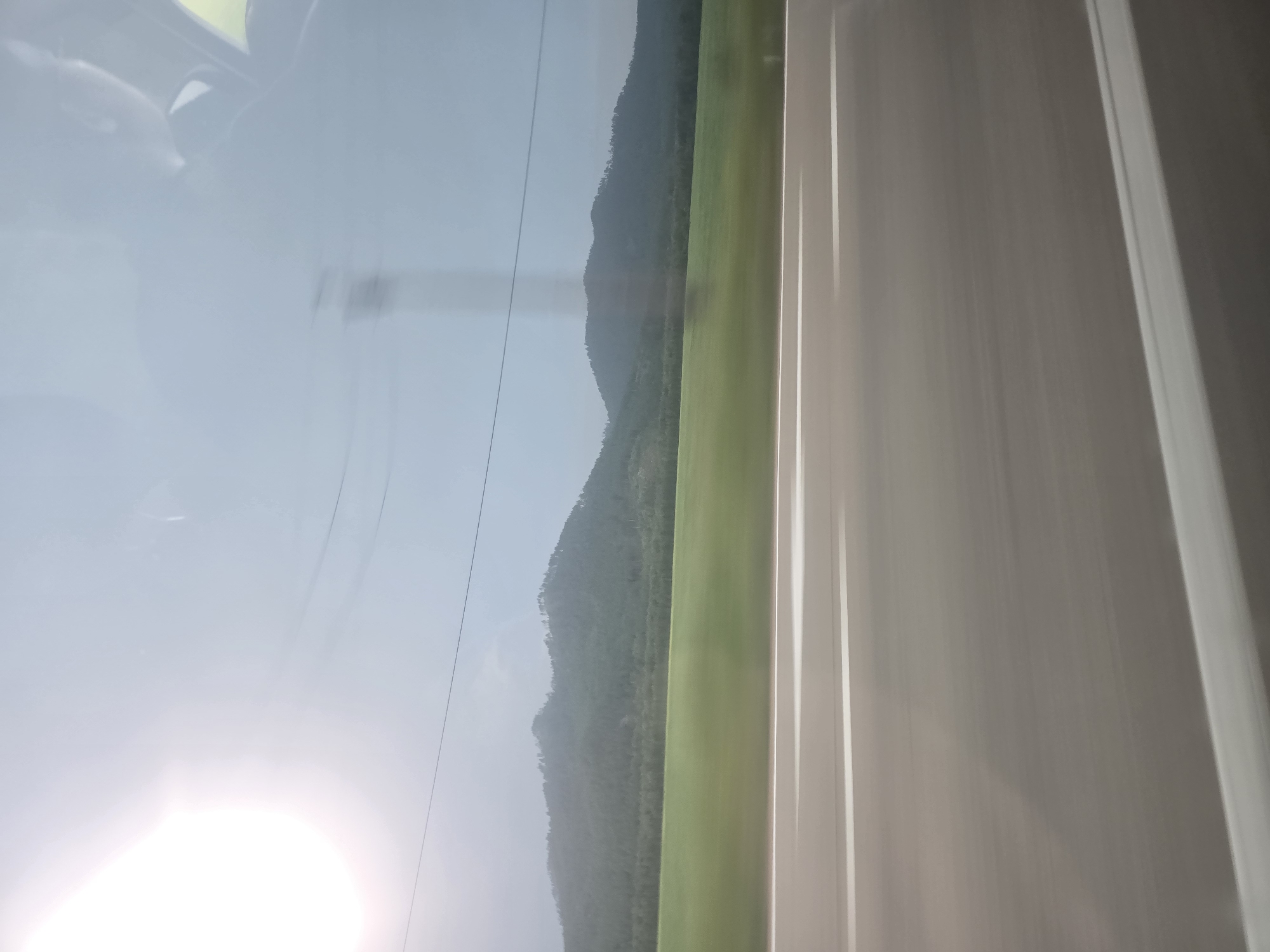 The image depicts a landscape seen from a moving vehicle, likely a car, on a sunny day. The foreground shows a blurred asphalt road with white lines, indicating motion. In the middle ground, there is a green field that transitions into a forested hillside with gently rolling mountains in the background. The sky is bright and appears slightly hazy, with the sun creating a lens flare in the upper left quadrant. There are no people or specific structures visible that would allow for precise identification of a city or location, therefore it is not possible to confirm the location as Baturino, Russia. The scene is typical of a rural or mountainous region viewed from a road.
