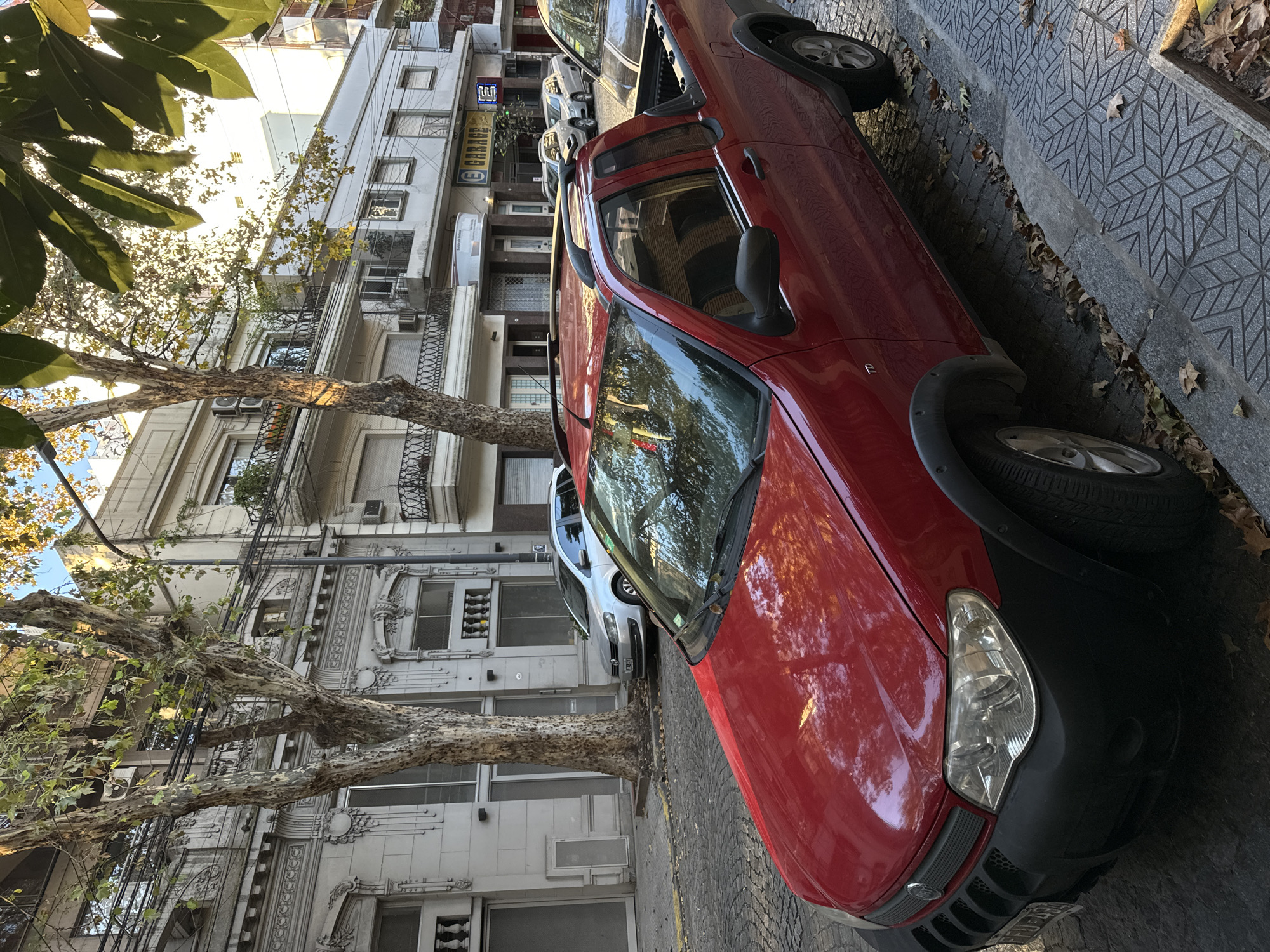 A red Fiat Strada pickup truck is parked on the right side of a street in Buenos Aires, Argentina. The street is paved with diamond-patterned concrete tiles. Buildings with balconies and ornate facades line the street. Trees with mottled bark and sparse foliage are present along the street, with branches extending into the view. Other parked vehicles, including a silver car, are visible further down the street. The lighting suggests it is daytime. The presence of dry leaves on the pavement indicates it may be autumn. A “Garage” sign can be seen on the buildings.