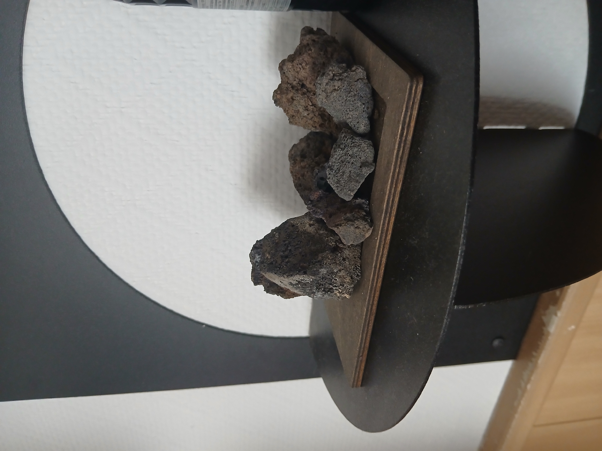 A cluster of dark, porous rocks is displayed on a small, dark wooden platform. The platform rests on a circular, black shelf, which is part of a larger black shelf unit attached to a white wall. The wall has a textured surface, possibly painted brickwork or stucco. A portion of a black, curved architectural element is visible behind the shelf and rocks. The lighting suggests an indoor environment. No location in Moscow, Russia is discernible from the image.