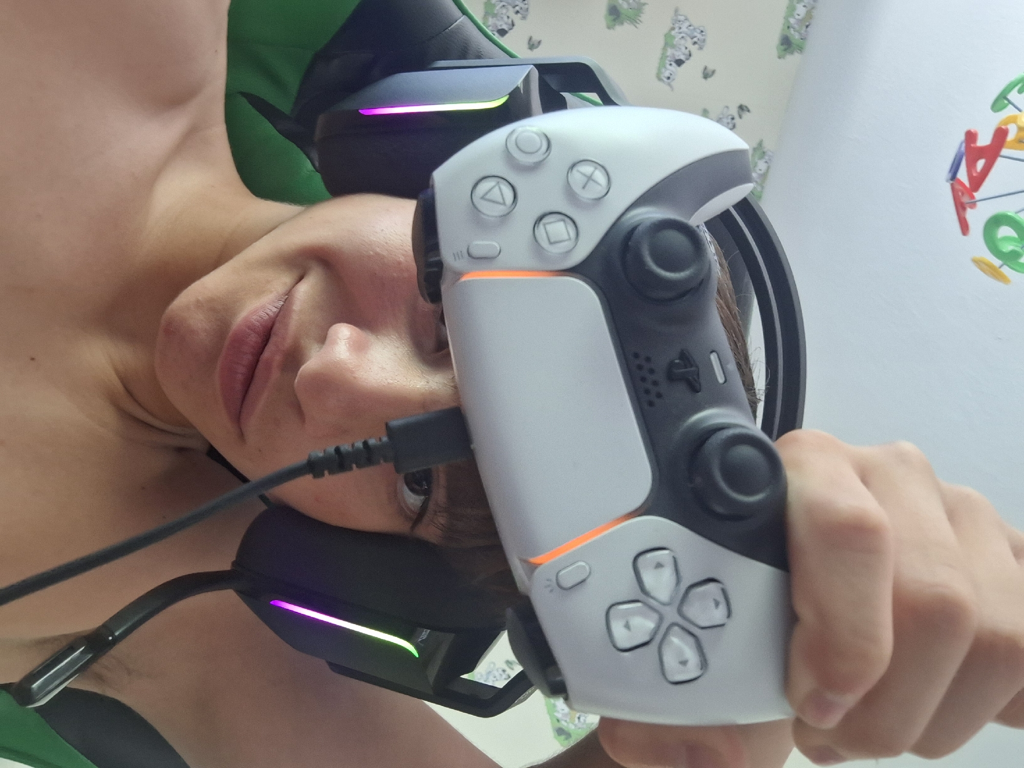 A young person, wearing gaming headphones with illuminated accents, holds a white and gray video game controller above their head. The controller, partially obscuring their face and forehead, is connected by a black cable. The background features a wall with patterned wallpaper depicting dogs and a mobile with colorful alphabet letters hanging from the ceiling. The scene appears to be an indoor personal space, possibly a bedroom. The image does not contain information that allows for the identification of its location in Varna, Bulgaria.