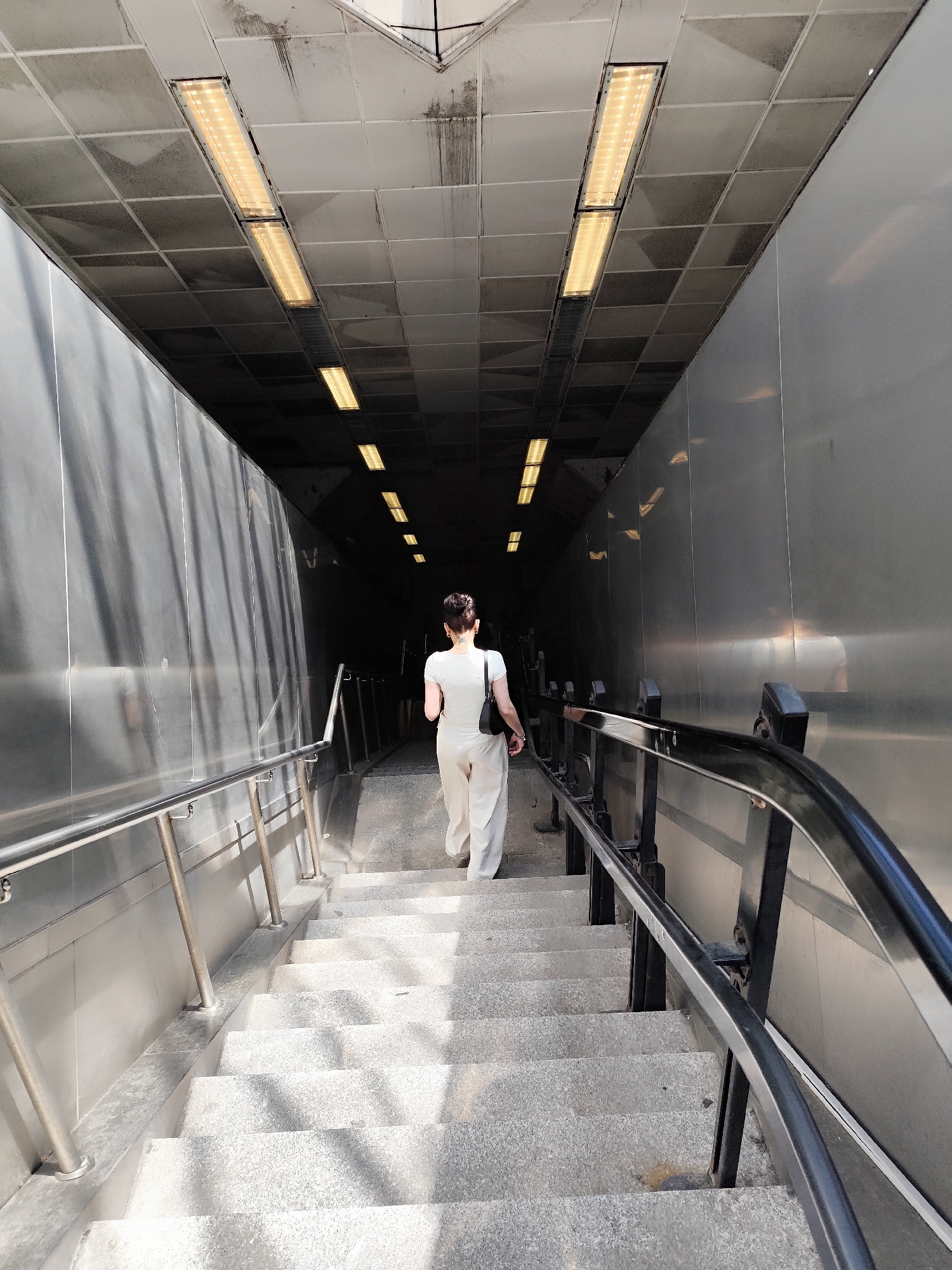 The image shows a woman walking down a staircase into an underground passageway or tunnel. The location is likely in Sarıyer, Turkey. The woman is wearing white clothing and carrying a black purse. The staircase is made of gray stone, and there are metal handrails on both sides. The tunnel ceiling is tiled, with evenly spaced fluorescent lights. The walls of the tunnel are a light gray metallic surface. The lighting conditions vary, with the entrance of the staircase being brightly lit, and the depth of the tunnel being darker.