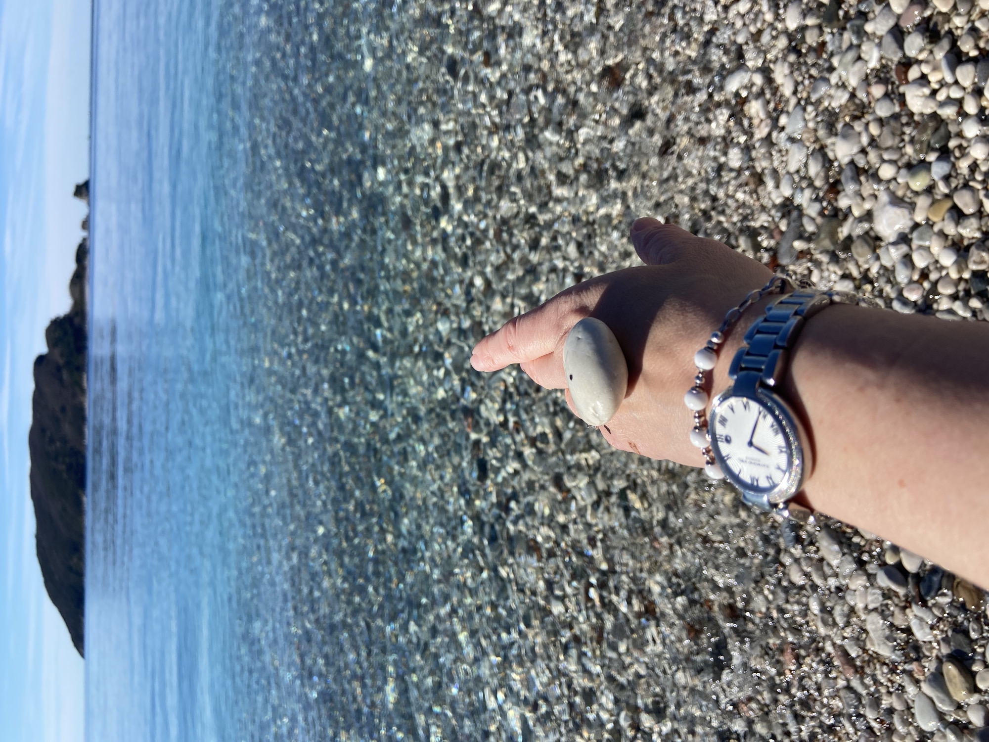 The image shows a hand holding a white, oval stone or shell on a beach in Budva, Montenegro. The background consists of clear, shallow water and a pebble beach. The hand is wearing a silver watch and a bracelet with white beads. In the distance, on the left, there is a blurry silhouette of land. The light suggests it is a sunny day.