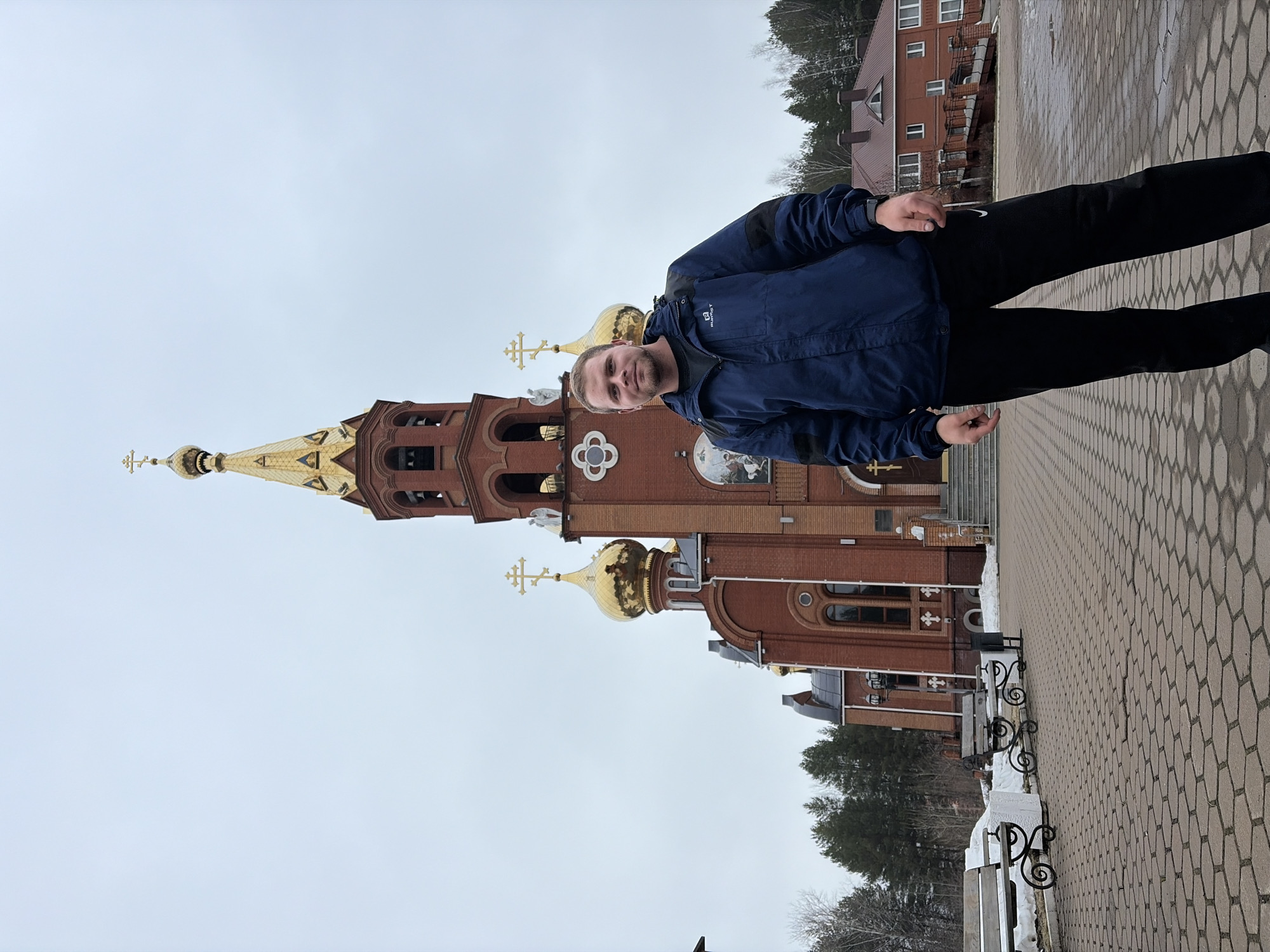 A medium shot shows a man standing on a brick-paved plaza with an Orthodox church in the background. The man is facing forward at a slight angle, looking at the camera. He has short blond hair and a beard. He wears a blue jacket and black pants.
The church is made of red brick with golden domes. The central dome is tall and pointed, while the smaller domes are more rounded. There are crosses atop each dome. The church has arched windows and decorative elements.
Behind the church is a two-story building, also made of brick, and some trees.
To the right of the man and the church, there are benches with decorative ironwork along the pathway. The sky is overcast. The location is likely the Orthodox Church in Chaikovsky, Russia.
