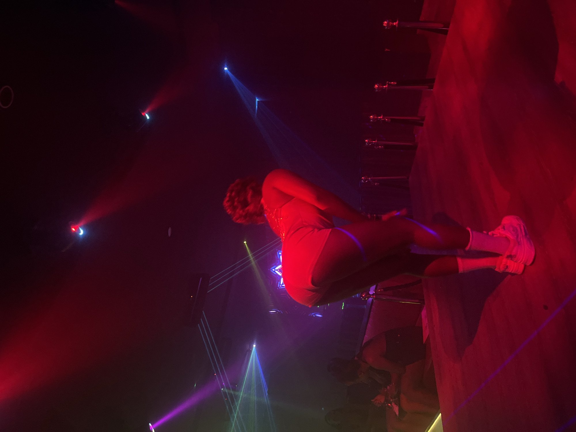 In a dimly lit nightclub setting, likely in Howayi, Nigeria, a woman is performing on a raised stage bathed in vibrant red and purple lights. She is bent over, her back to the camera, wearing white shorts and white socks with chunky white sneakers. The stage is covered in a dark red carpet, and a velvet rope stanchion line is visible in the foreground. Laser lights crisscross the background, illuminating the smoky air. In the background, other patrons are seated at tables. The overall atmosphere suggests a lively entertainment venue.