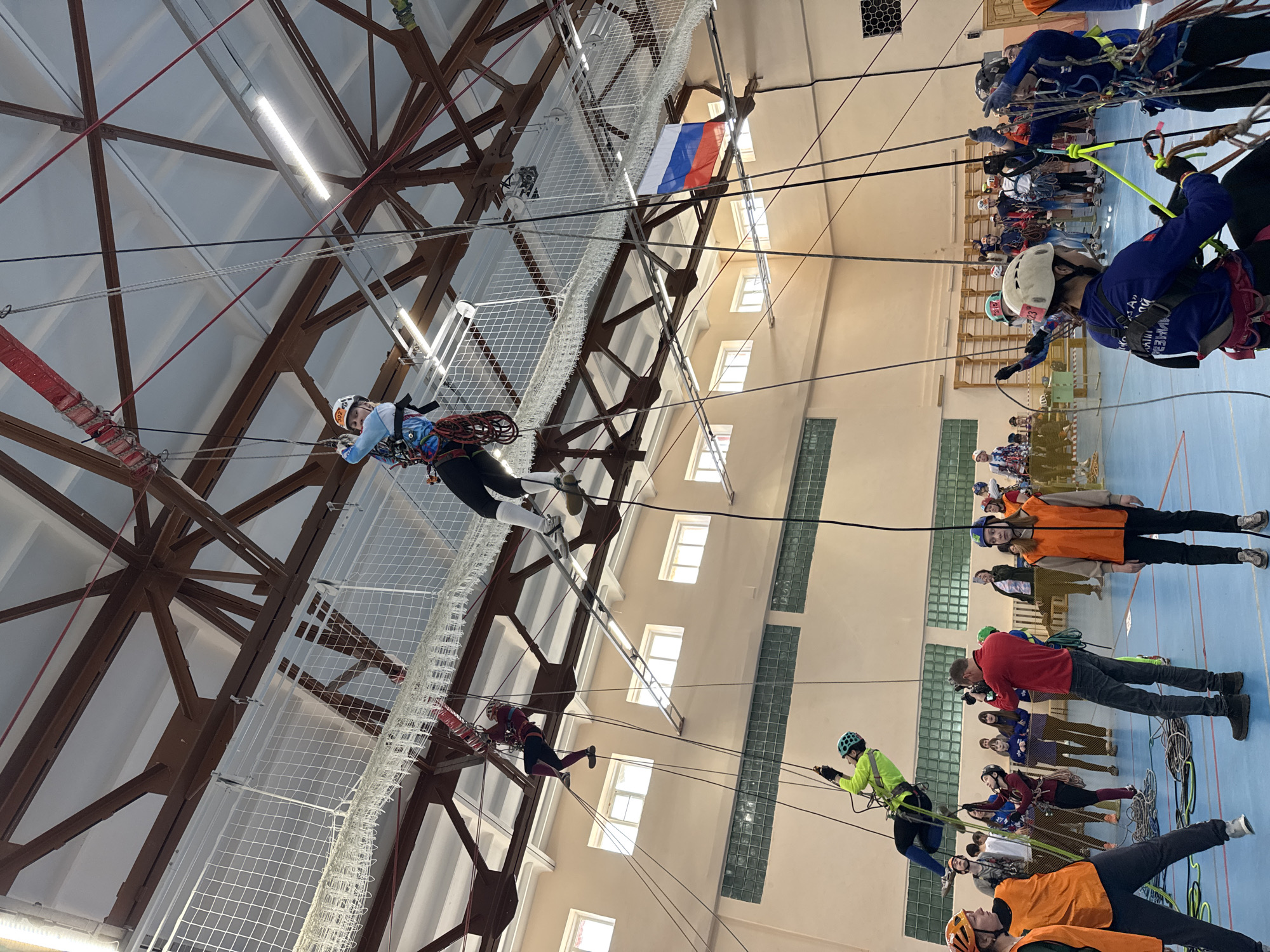 The image depicts an indoor climbing competition in Usolye, Russia. Participants wearing helmets and harnesses are suspended from ropes, navigating a course constructed from metal beams and netting beneath the building's ceiling. Spectators, some wearing orange vests, stand below observing the event. A Russian flag hangs near the climbing structure.