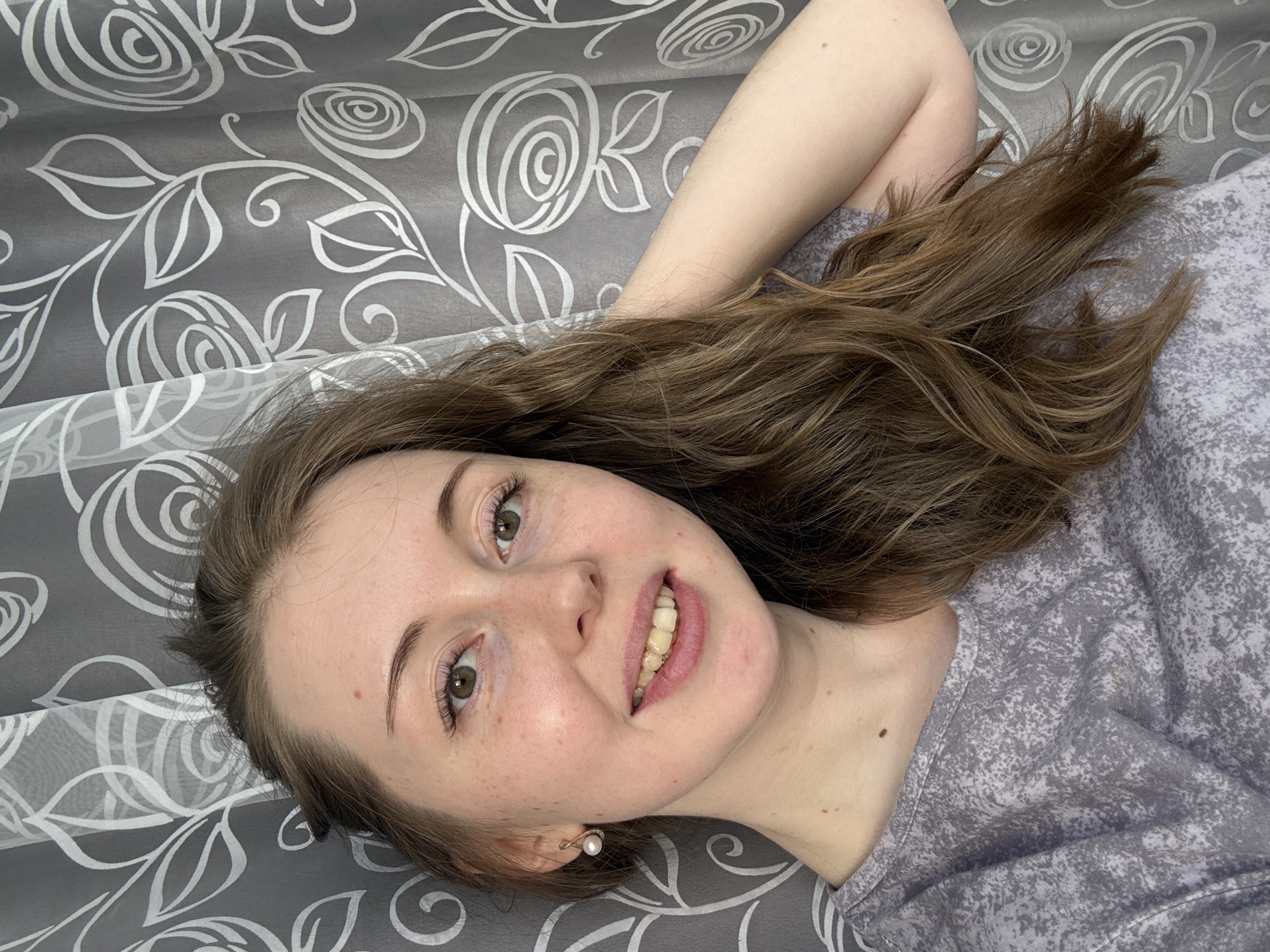 A young woman is lying on a gray patterned surface. Her head is tilted slightly, and she is looking directly at the viewer with a smile. Her hair is brown and styled loosely around her face. She is wearing a gray patterned shirt. An earring is visible on her left ear. Her left arm is bent and positioned behind her head. The location is Unknown, Unknown.