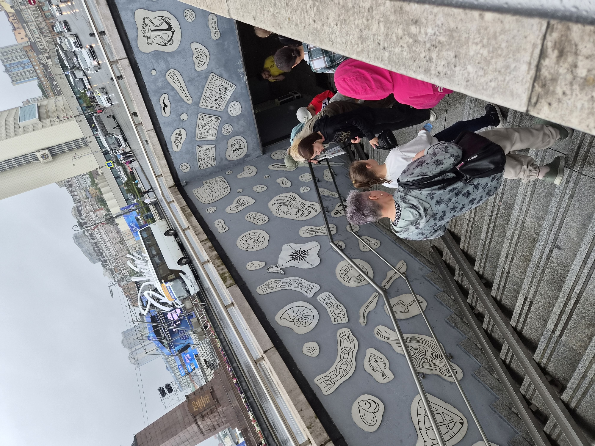 The image depicts a group of people descending a wide, grey stone staircase into a pedestrian underpass. The underpass walls are decorated with numerous white, stylized reliefs of marine life and nautical symbols, such as shells, fish, chains, anchors, and compasses. A metal handrail runs along the side of the stairs.
In the background, above the underpass entrance, a busy city street is visible. Cars, buses, and pedestrians are present. A white tour bus with illuminated signage is prominently featured, suggesting a tourist attraction or event. The urban landscape includes various buildings, some modern and tall, others older and ornate, characteristic of Vladivostok, Russia. The sky is overcast, indicating a cloudy or potentially rainy day. The overall scene captures a moment in a public urban space, highlighting both architectural details and everyday city life.