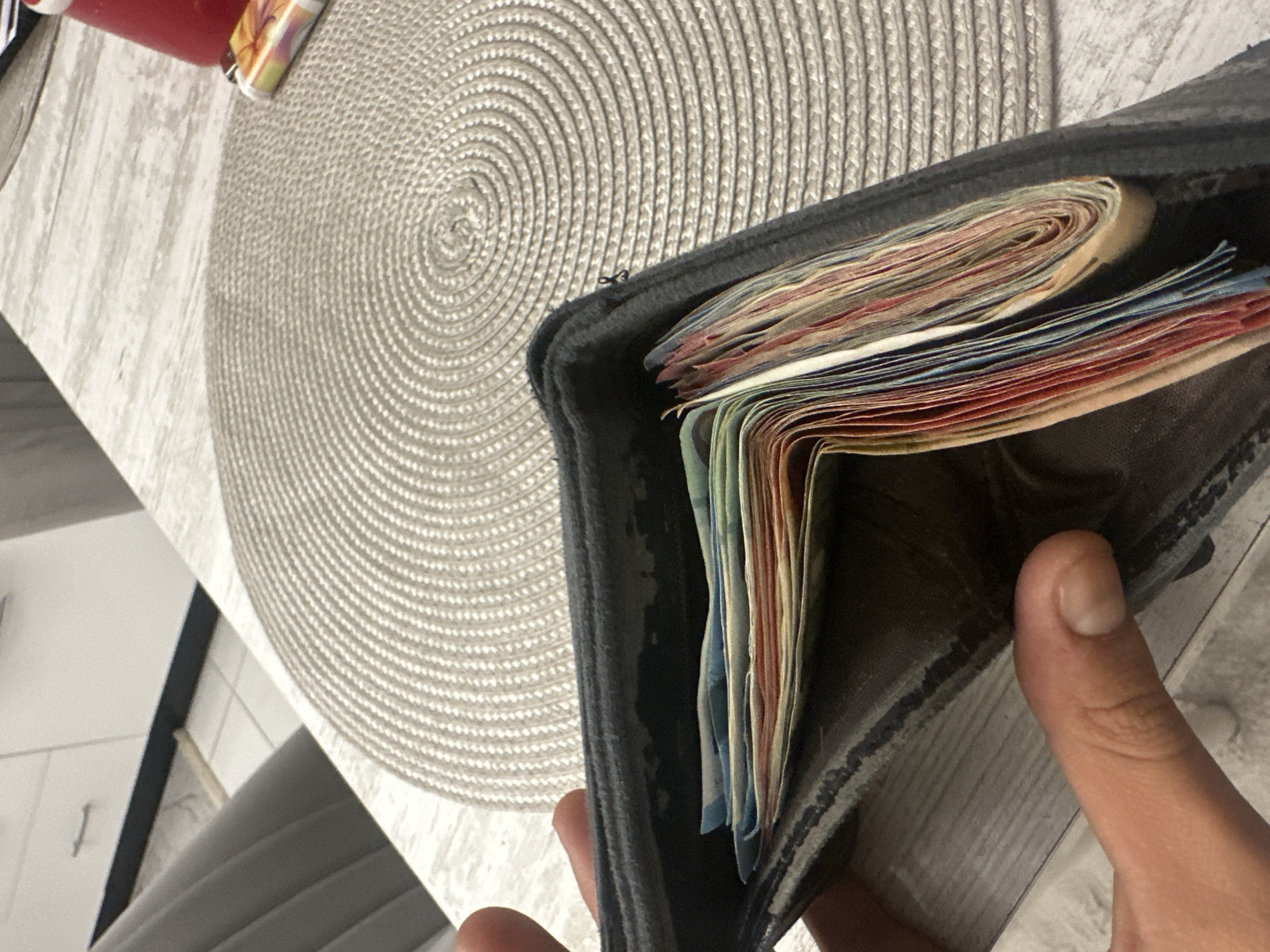 An indoor scene, reported to be located in Sofia, Bulgaria, captures a close-up perspective of a hand holding an open, dark-colored wallet. The wallet is substantially filled with numerous folded banknotes of varying colors, including shades of blue, red, green, and yellow, indicating a significant quantity of currency. The hand, exhibiting a light to medium skin tone, is positioned in the lower left portion of the frame, with the thumb and part of the index finger visible. The wallet rests partially on a light-colored table or countertop that features a wood-grain texture. A circular, light beige or gray placemat with a tightly woven, concentric spiral pattern occupies a large portion of the table surface in the midground. To the right on the placemat, a red cylindrical object, possibly a cup or bottle, and a disposable lighter with a multi-colored, abstract design are visible. In the background, to the left, a gray upholstered chair with vertical ribbing is partially visible. Further back, to the right, white kitchen cabinets with a single silver horizontal handle are discernible, above a white tiled floor with dark grout lines and a dark baseboard or kickplate. No other individuals or active interactions are depicted.