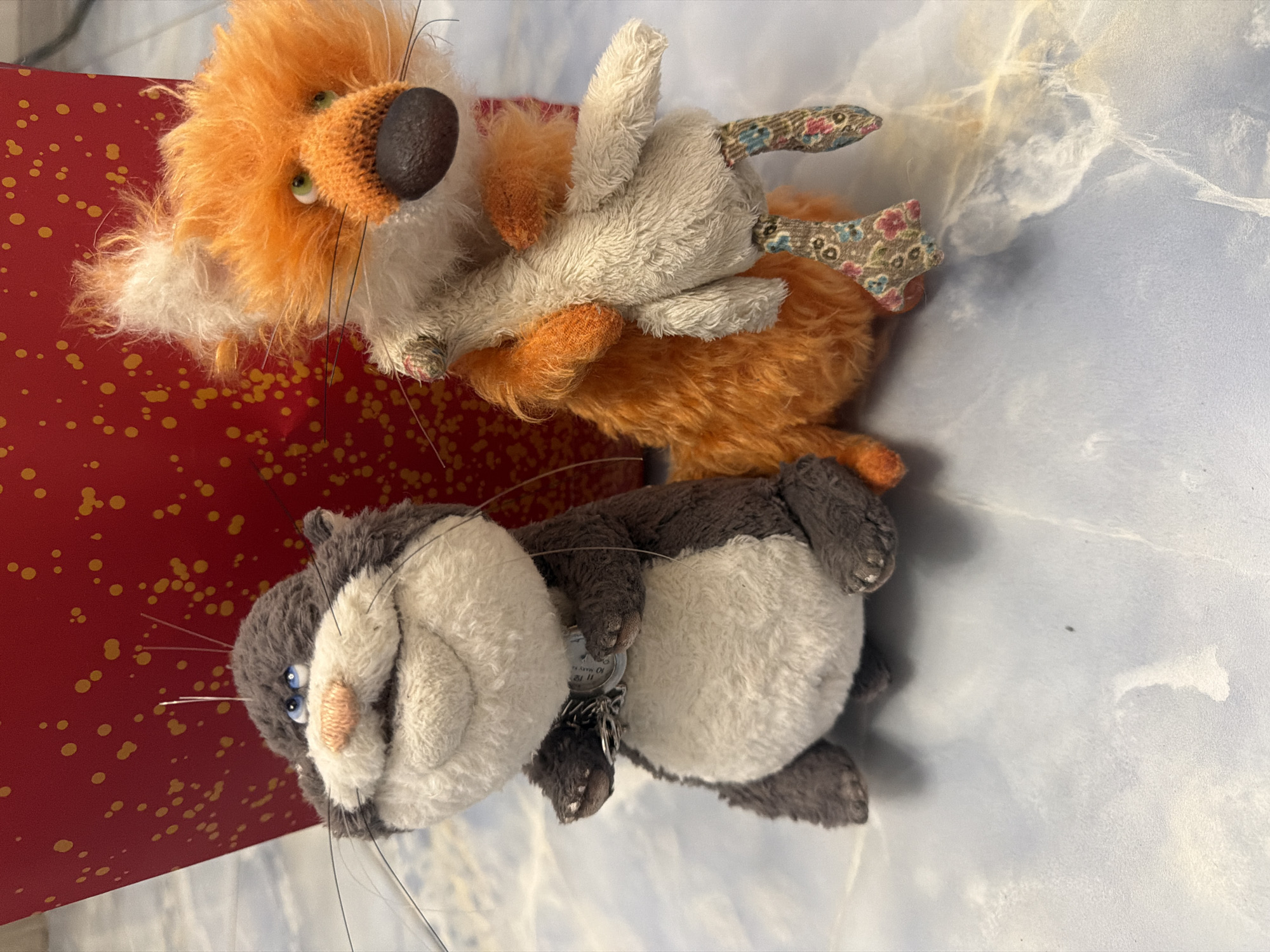 Two stuffed animals, a gray and white cat and an orange fox, are positioned on a light blue surface patterned with white and gray swirls. The cat, on the left, is facing slightly to the right and has blue eyes, a pink nose, and whiskers. It wears a silver chain with a circular tag around its neck. The fox, on the right, is facing forward and is holding a light-colored stuffed animal in its arms. The fox has green eyes, a dark brown nose, and a fluffy orange and white body. Both animals are in focus and appear to be sitting. Behind the animals is a large, out-of-focus red object with scattered yellow dots. The lighting is bright and appears to come from above. The scene is static with no visible motion.