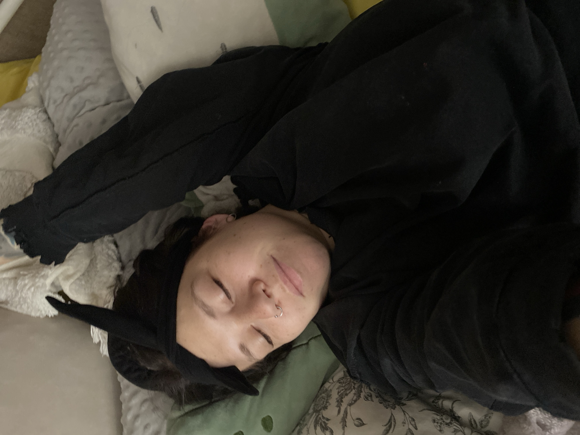 A young woman with fair skin is lying down, eyes closed, on a bed or similar surface. She wears a black hoodie and a black headband with pointed ears. A silver ring is visible in her left nostril. The bedding includes various textures and colors, including gray, white, green, and yellow. The background appears blurry. The location is unknown and there is no information to indicate that it is in Chaikovsky, Russia.