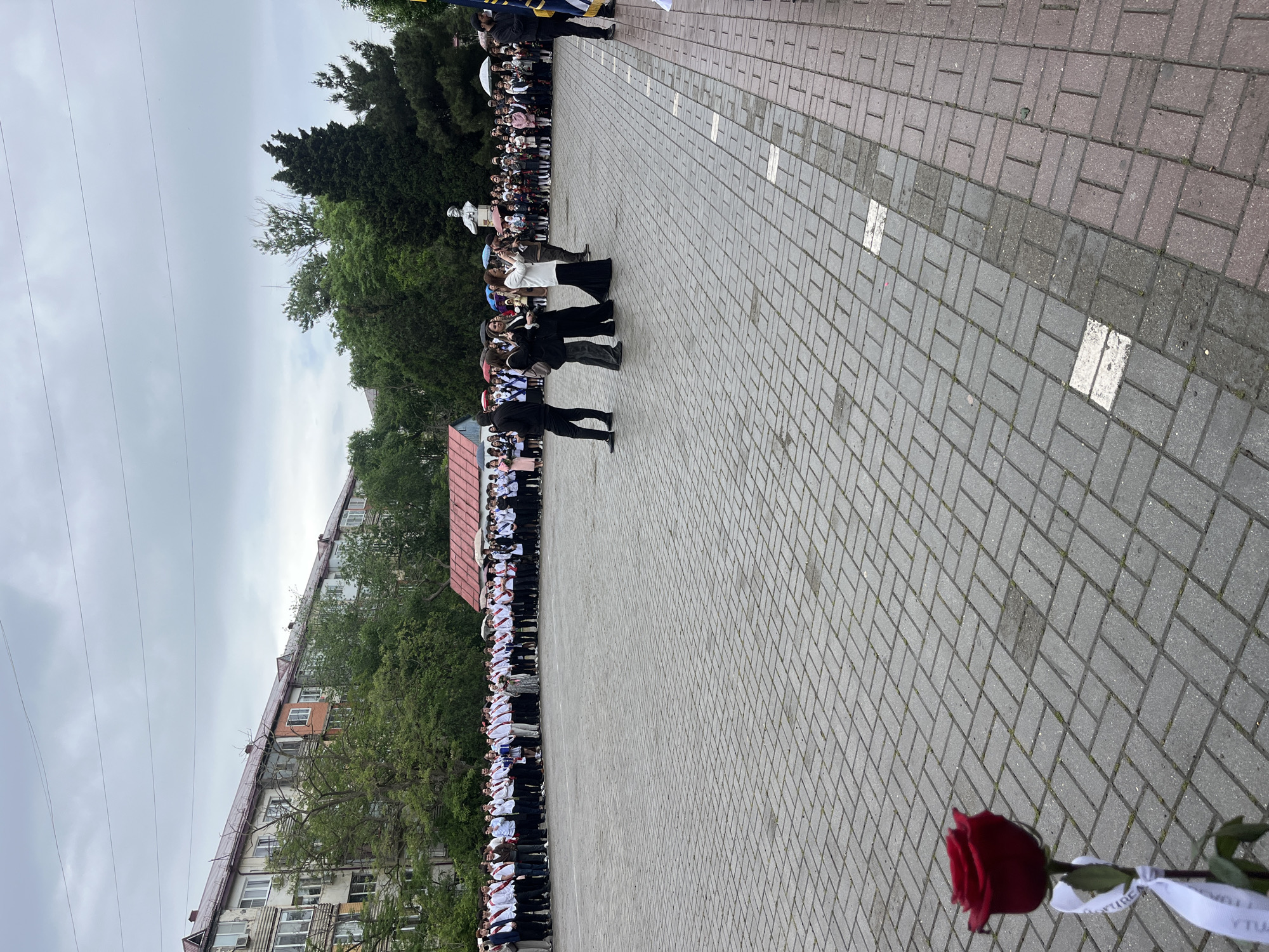 The image captures an outdoor event on a brick-paved square. A line of people stands to the left, seemingly in formal attire with many wearing white shirts or blouses. A building is visible in the background, with trees interspersed. To the right, a red rose with a white ribbon lies on the pavement. The scene gives the impression of a formal gathering, possibly a ceremony or school event. Kaspiysk, Russia, based on the ribbon.